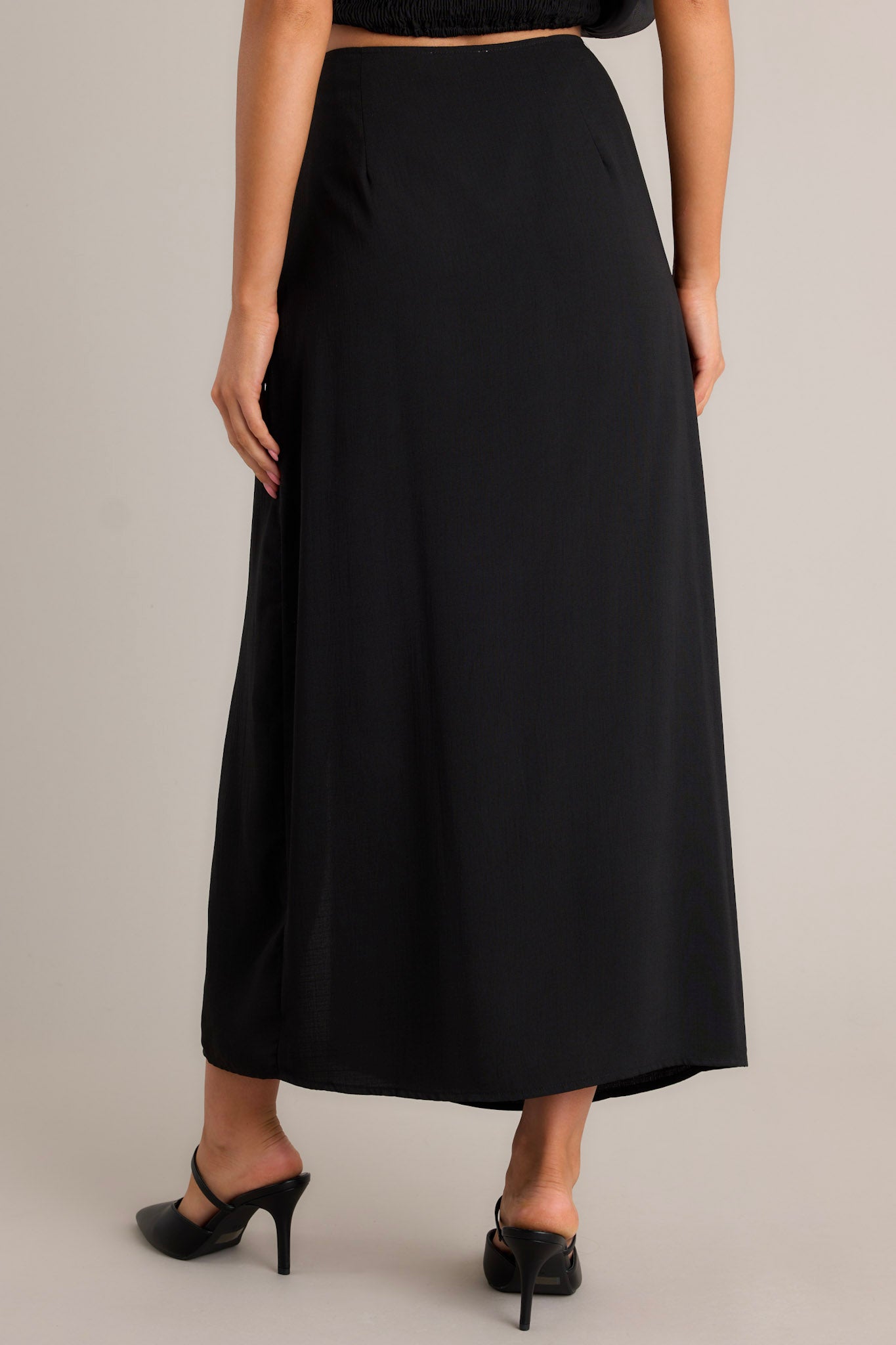 Back view of this black midi skirt featuring a high waisted design, pleated detailing on the hip, faux buttons, a discrete side zipper, and a side slit.
