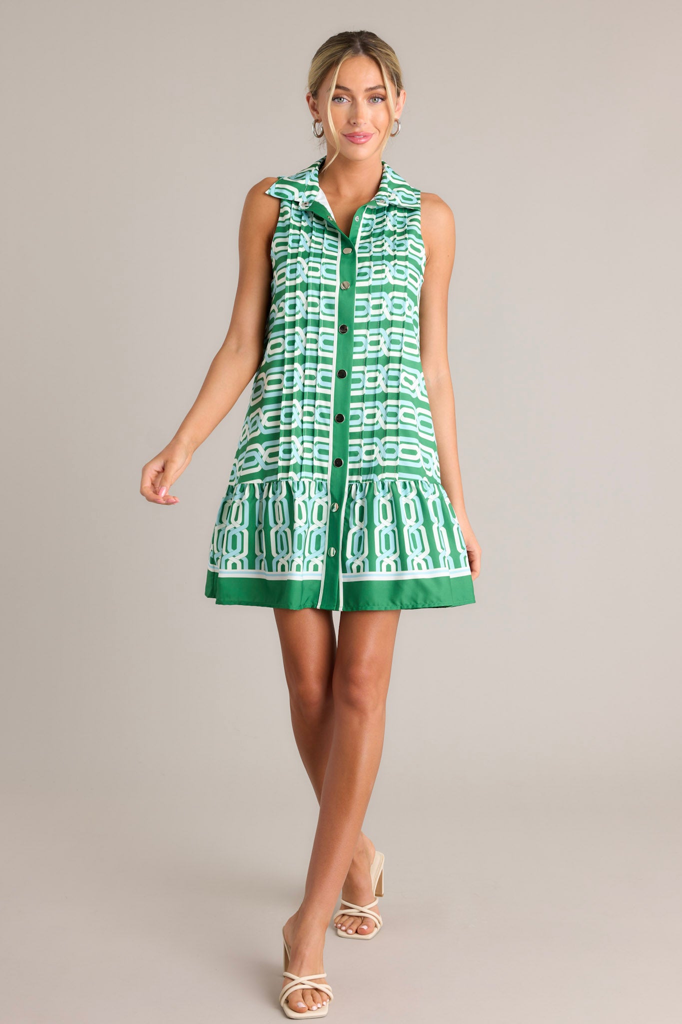 Full length view of a green multi mini dress with a collared neckline, a functional button front, pleats, a unique pattern, a single tier, and a sleeveless design