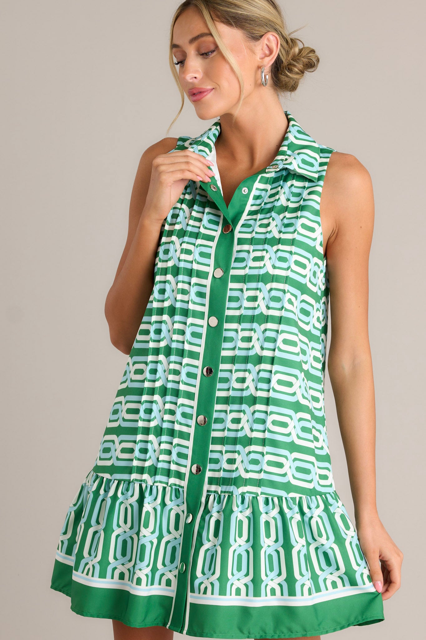 Front angled view of a green multi mini dress featuring a collared neckline, a functional button front, pleats, a unique pattern, a single tier, and a sleeveless design