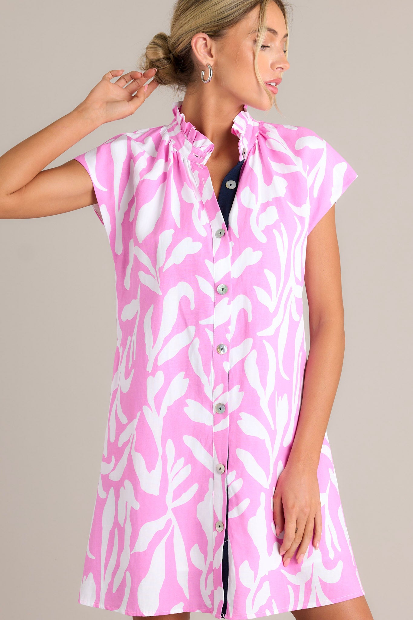 Front view of a pink dress featuring a ruffled collar, caped short sleeves, functional buttons, a fun abstract print, and functional hip pockets.