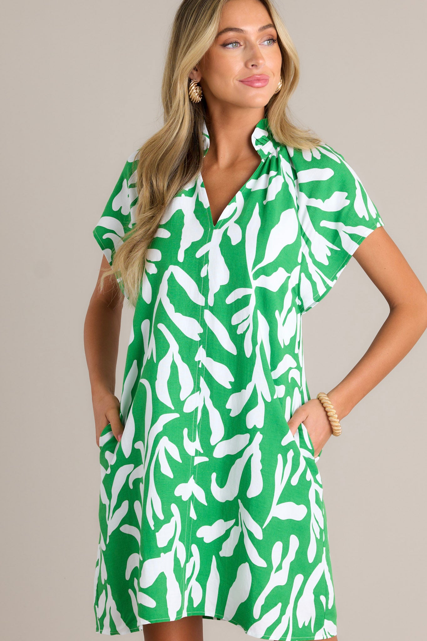 Front angled view of a green floral mini dress featuring a ruffled v-neckline, a unique floral design, functional hip pockets, and short flutter sleeves