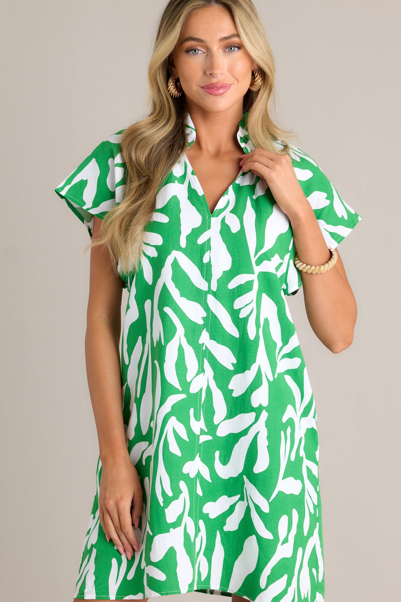 Front view of a green floral mini dress featuring a ruffled v-neckline, a unique floral design, functional hip pockets, and short flutter sleeves.