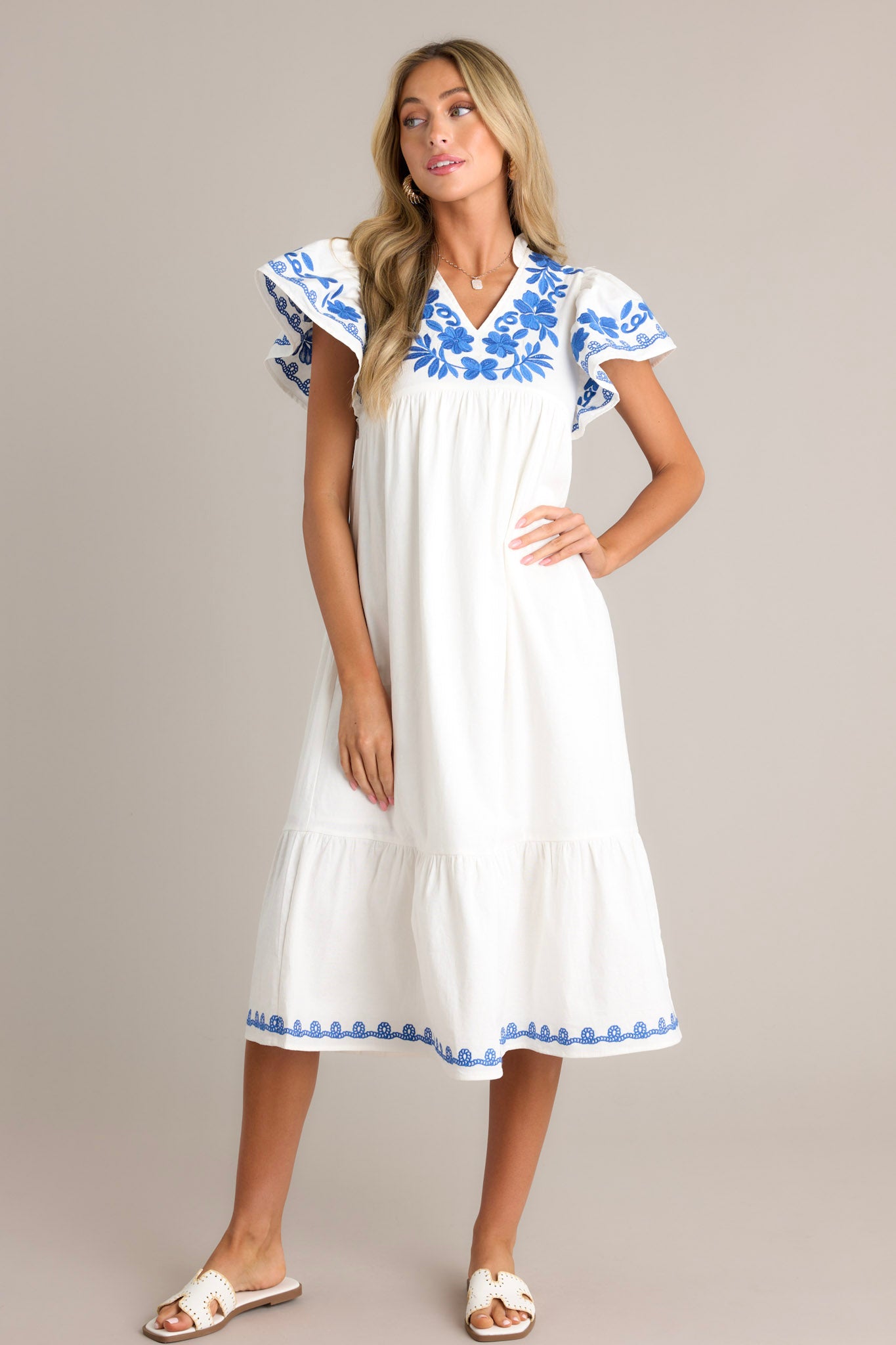 Front angled view of an ivory midi dress featuring a ruffled v-neckline, blue floral embroidery, a single tier, an embroidered hemline, and short embroidered flutter sleeves