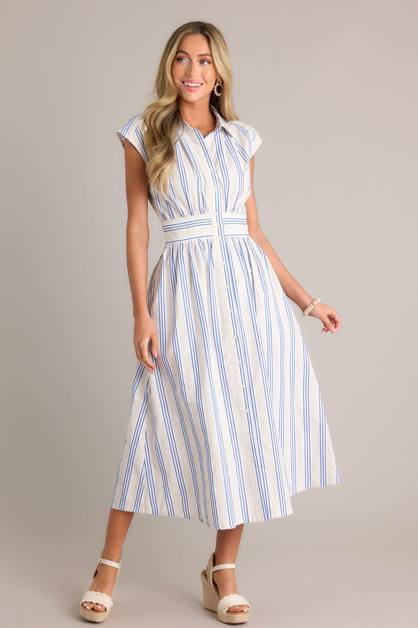 Action shot of a blue stripe midi dress displaying the fit and movement, highlighting the collared neckline, functional button front, functional hip pockets, beige and blue stripe pattern, and flowing silhouette.