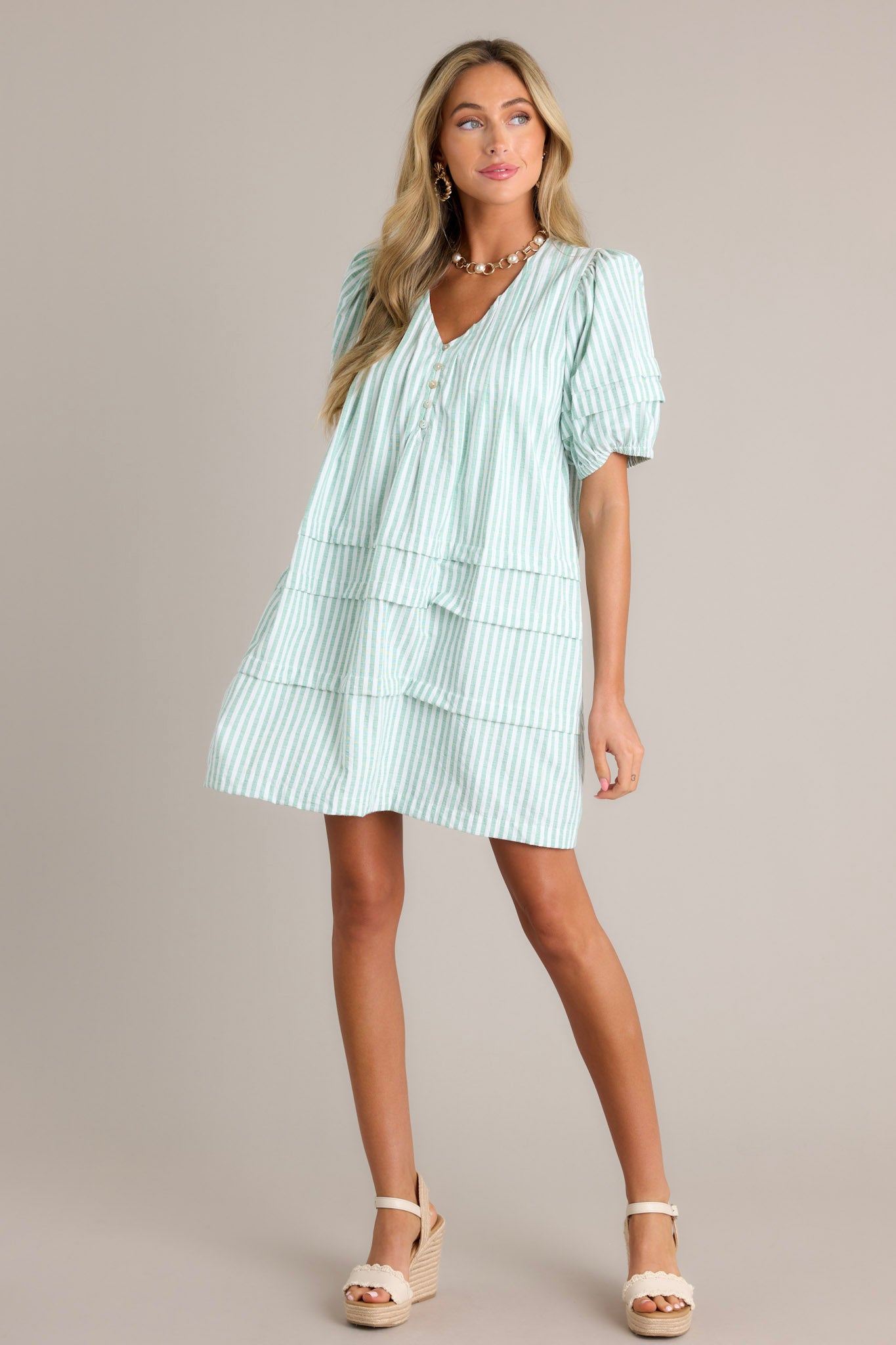 Front angled view of a mint green mini dress featuring a v-neckline, slight shoulder enhancement, functional buttons, subtle chest pleats, functional hip pockets, a tiered design, and elastic cuffed puff sleeves