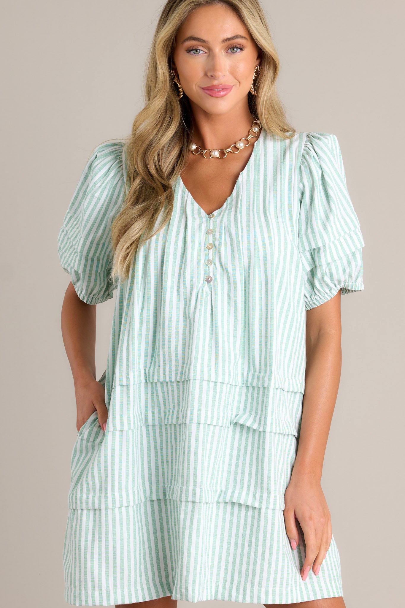 Front view of a mint green mini dress featuring a v-neckline, slight shoulder enhancement, functional buttons, subtle chest pleats, functional hip pockets, a tiered design, and elastic cuffed puff sleeves.