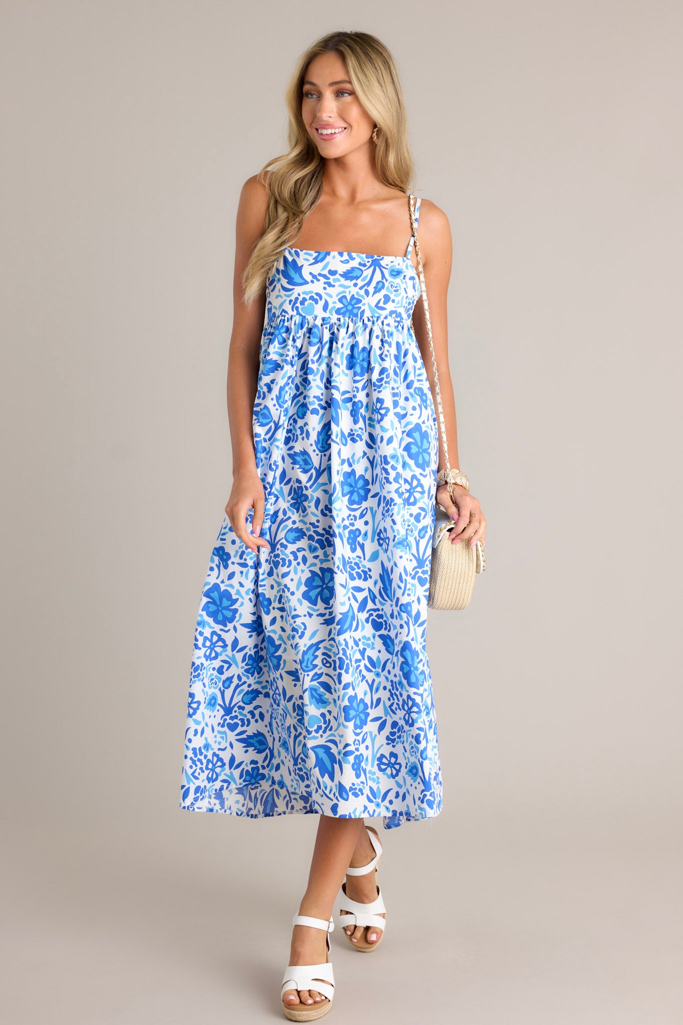 This blue floral maxi dress features a square neckline, thin adjustable straps, a self-tie back feature, a small cutout detail, functional hip pockets, and a flowing silhouette.