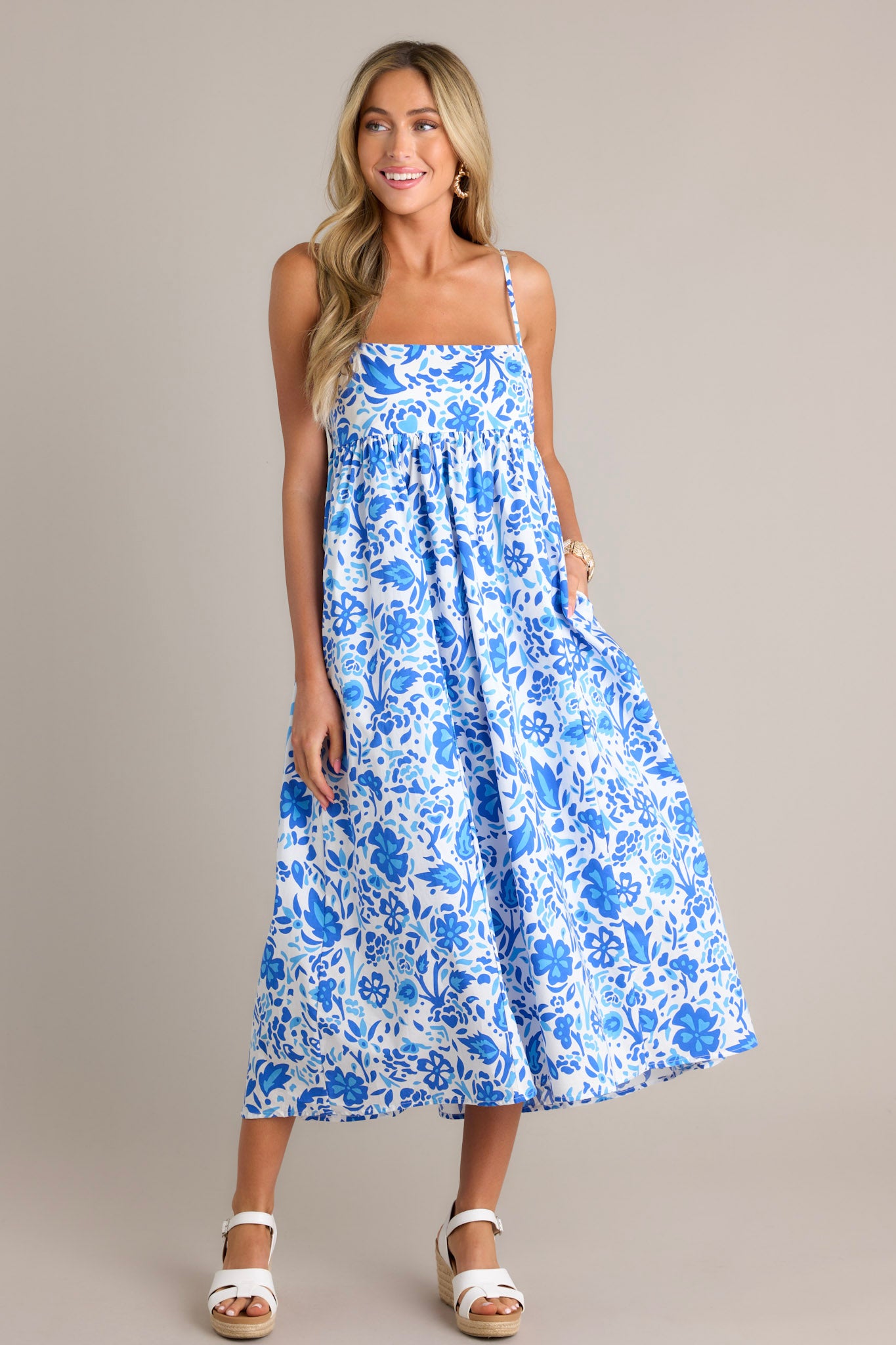 Full length view of a blue floral maxi dress with a square neckline, thin adjustable straps, a self-tie back feature, a small cutout detail, functional hip pockets, and a flowing silhouette