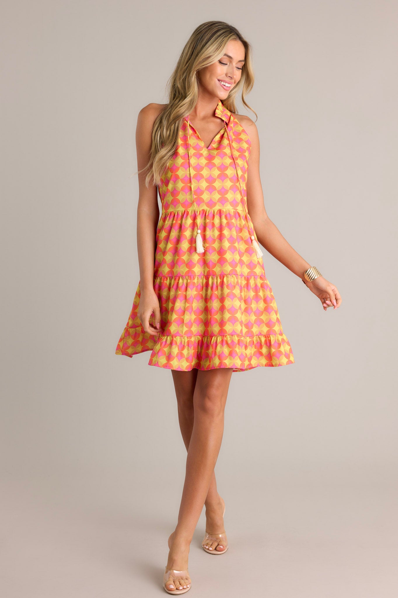 Front angled view of an orange mini dress featuring a ruffled v-neckline, a self-tie neck feature, a unique pattern, functional hip pockets, multiple tiers, and a flowing silhouette