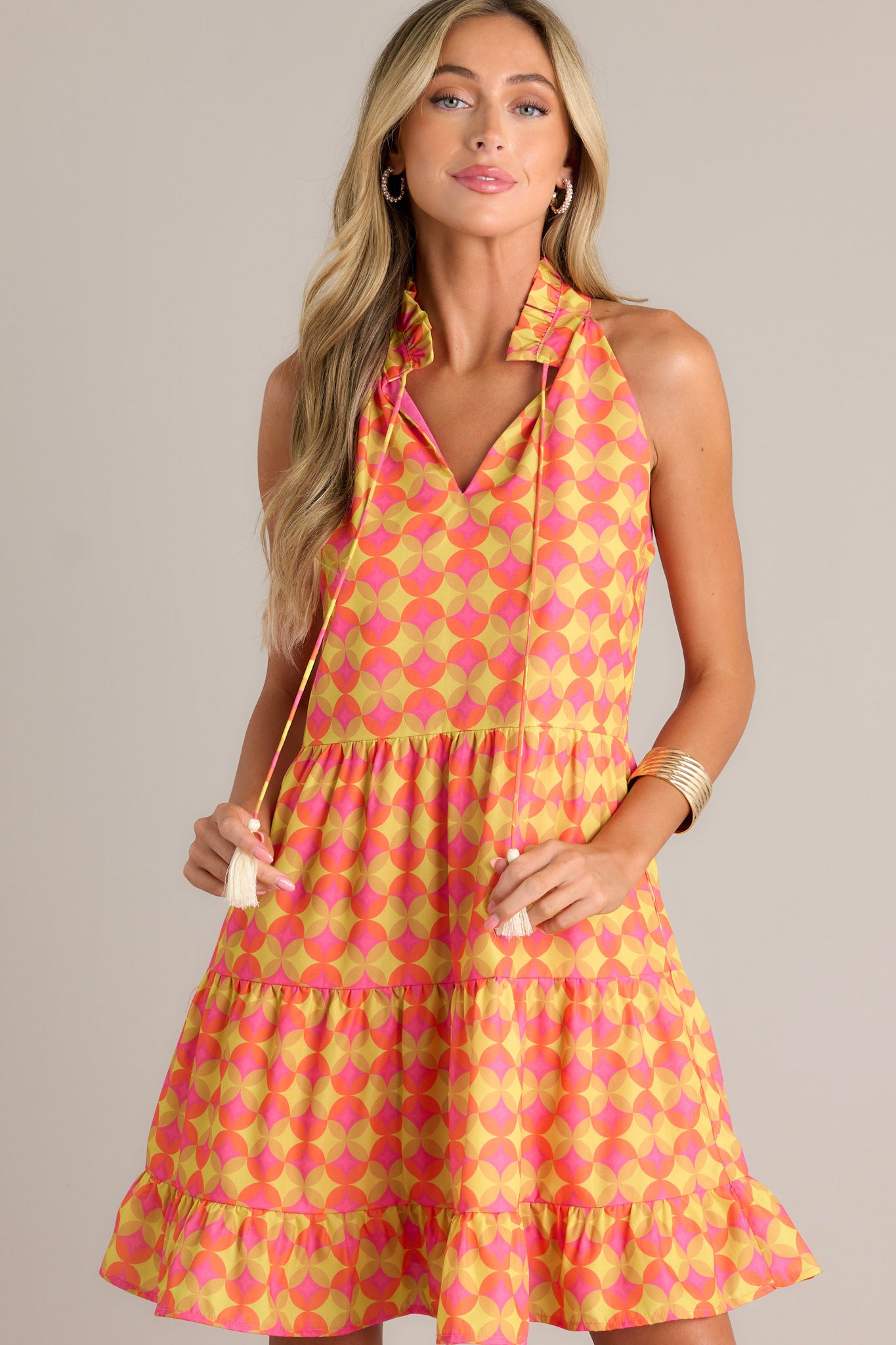 This orange mini dress features a ruffled v-neckline, a self-tie neck feature, a unique pattern, functional hip pockets, multiple tiers, and a flowing silhouette.