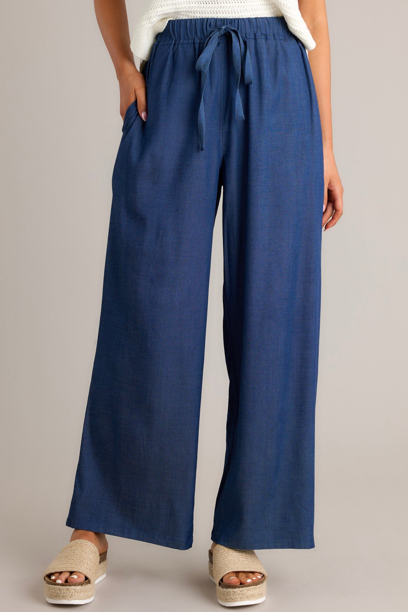 Front view of dark chambray pants featuring a high waisted design, an elastic waistband, a self-tie drawstring, functional hip pockets, and a wide leg.