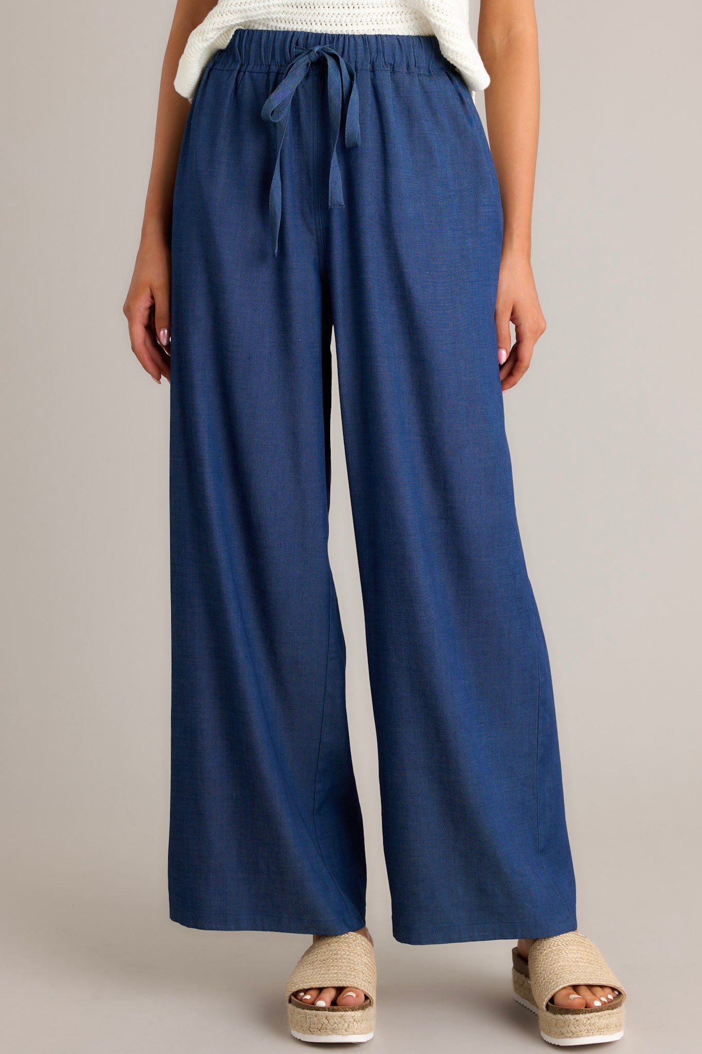 Full length view of dark chambray pants with a high waisted design, an elastic waistband, a self-tie drawstring, functional hip pockets, and a wide leg