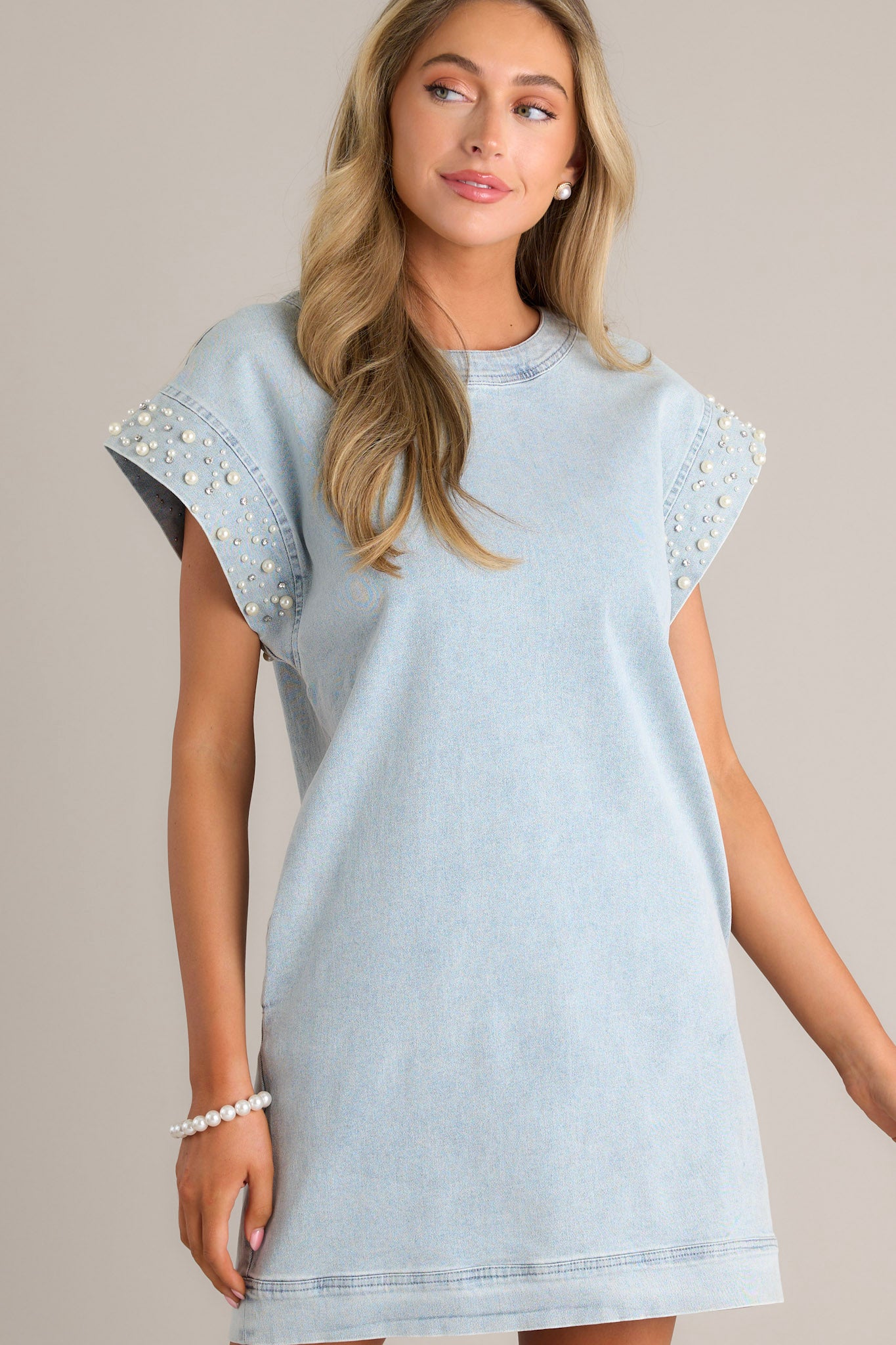 Front angled view of a light wash mini dress featuring a rounded neckline, denim material, functional hip pockets, and wide cap sleeves with pearl & rhinestone embellishments