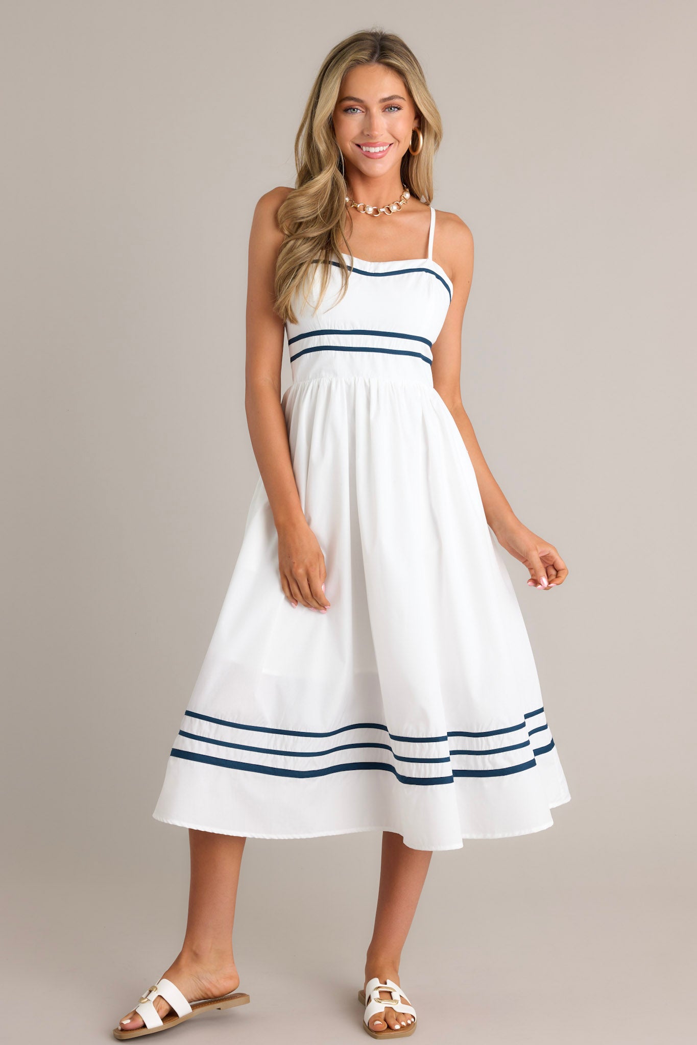 Front view of an ivory midi dress featuring a slight sweetheart neckline, navy trim, thin adjustable straps, a smocked back insert, a navy striped waist, functional hip pockets, and a navy striped hemline.