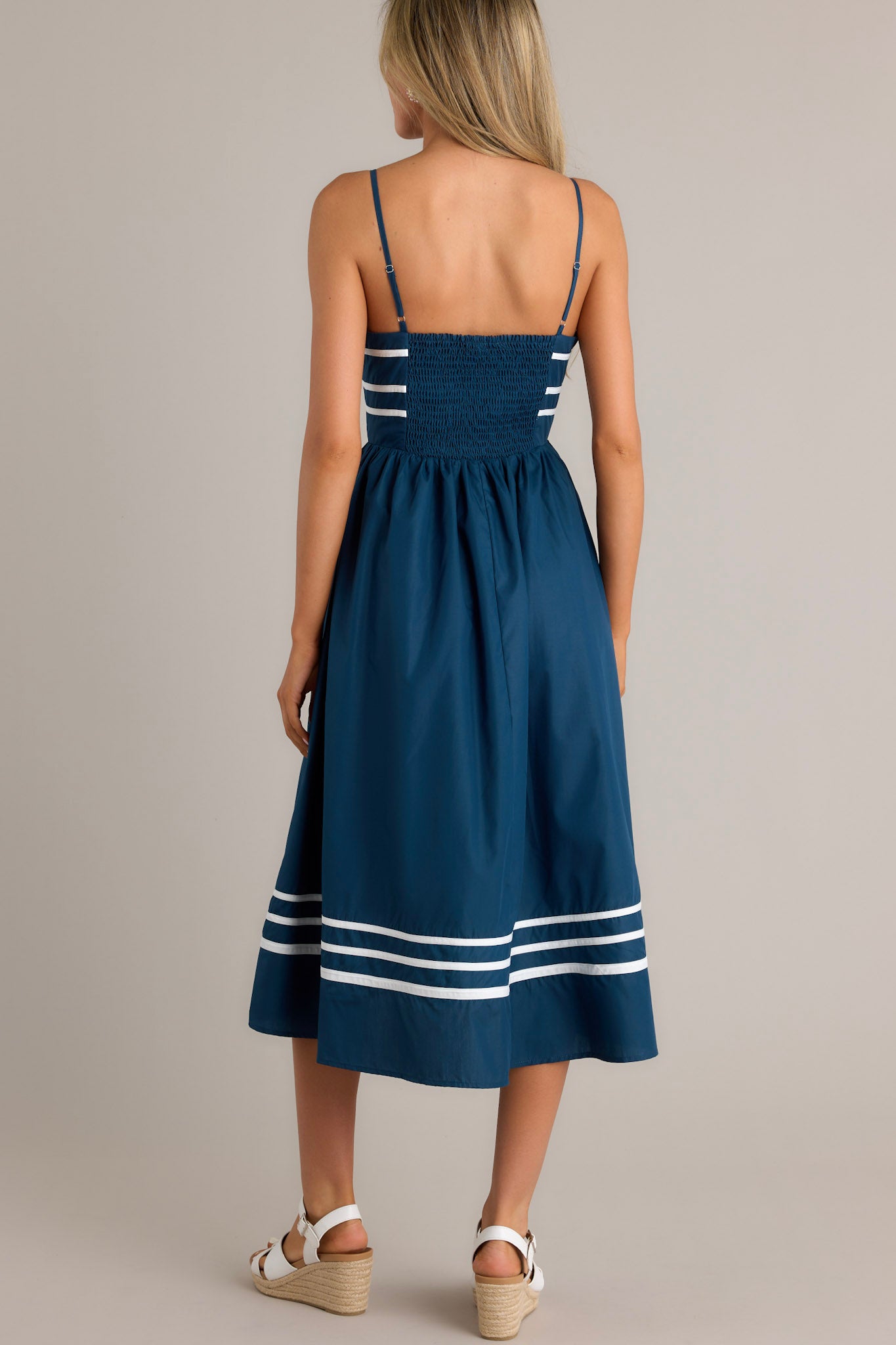 Back view of a navy midi dress highlighting the smocked back insert, thin adjustable straps, and an ivory striped hemline.