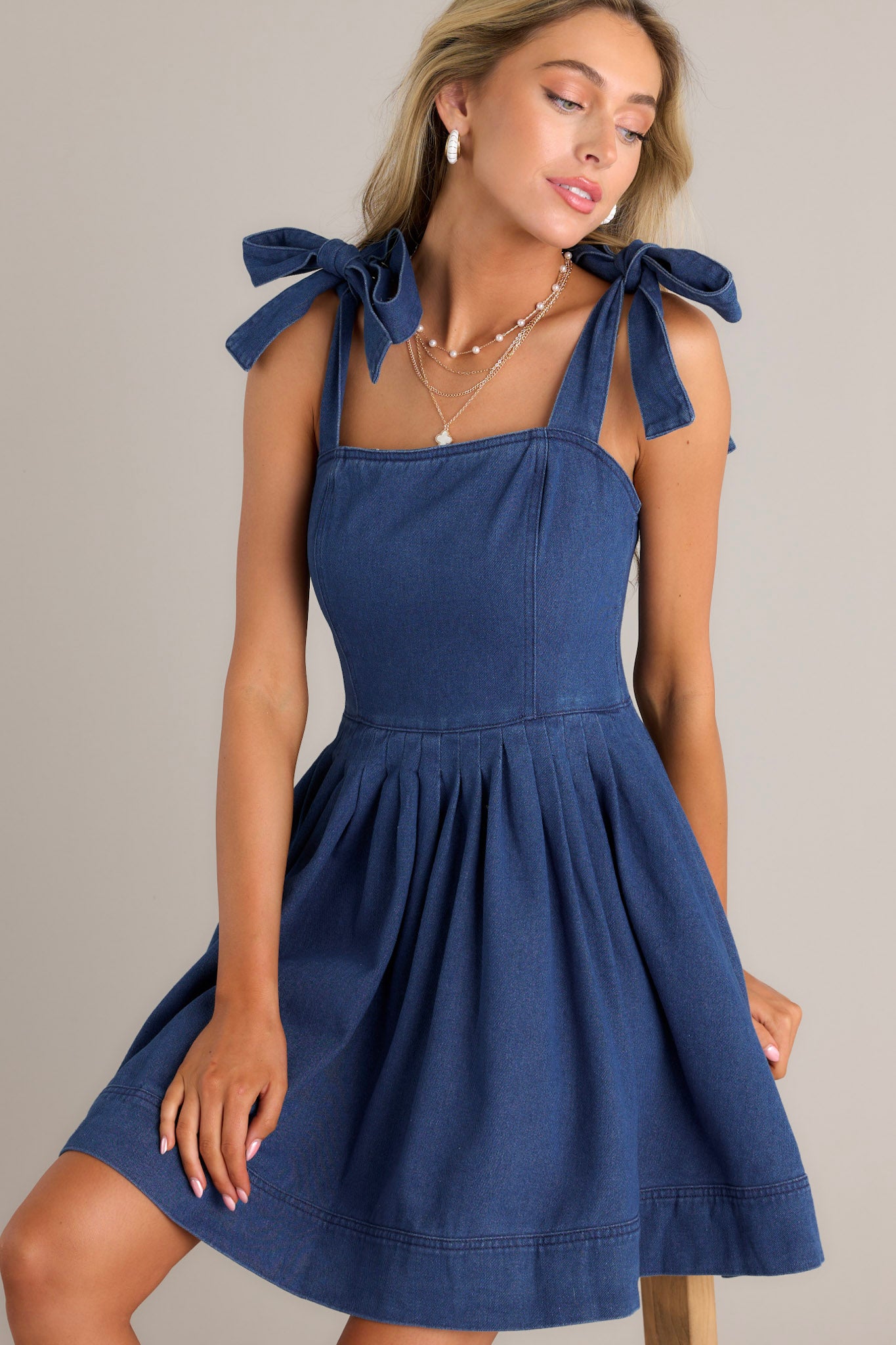 Front angled view of a denim mini dress featuring a square neckline, thick self-tie straps, subtle pleats throughout, functional hip pockets, and a thick hemline