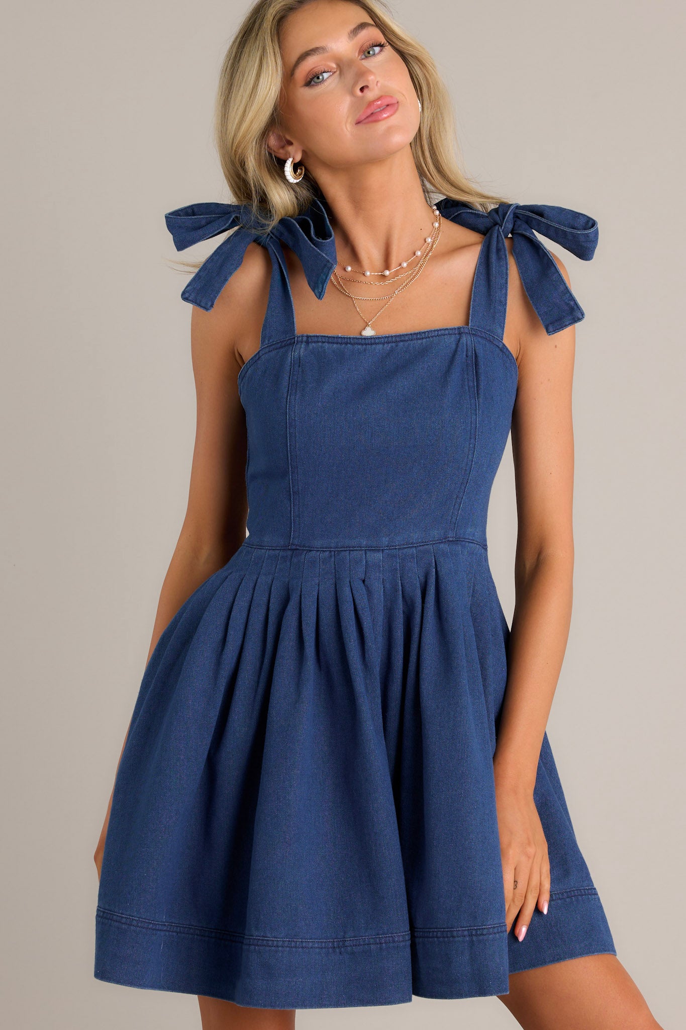 Front view of a denim mini dress featuring a square neckline, thick self-tie straps, subtle pleats throughout, functional hip pockets, and a thick hemline.