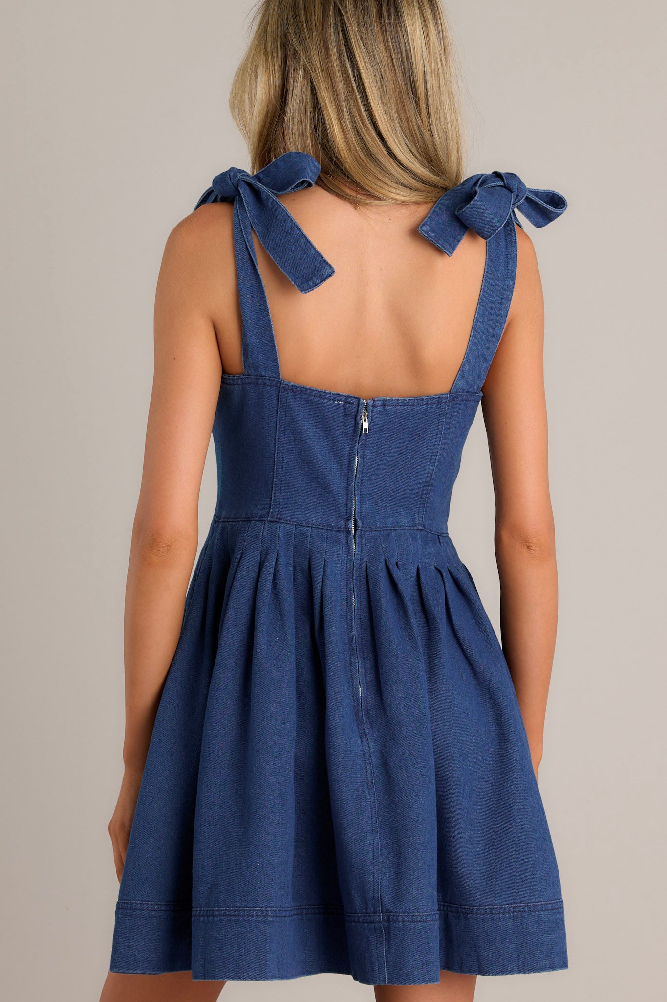 Back view of a denim mini dress highlighting the back zipper, thick self-tie straps, and subtle pleats.