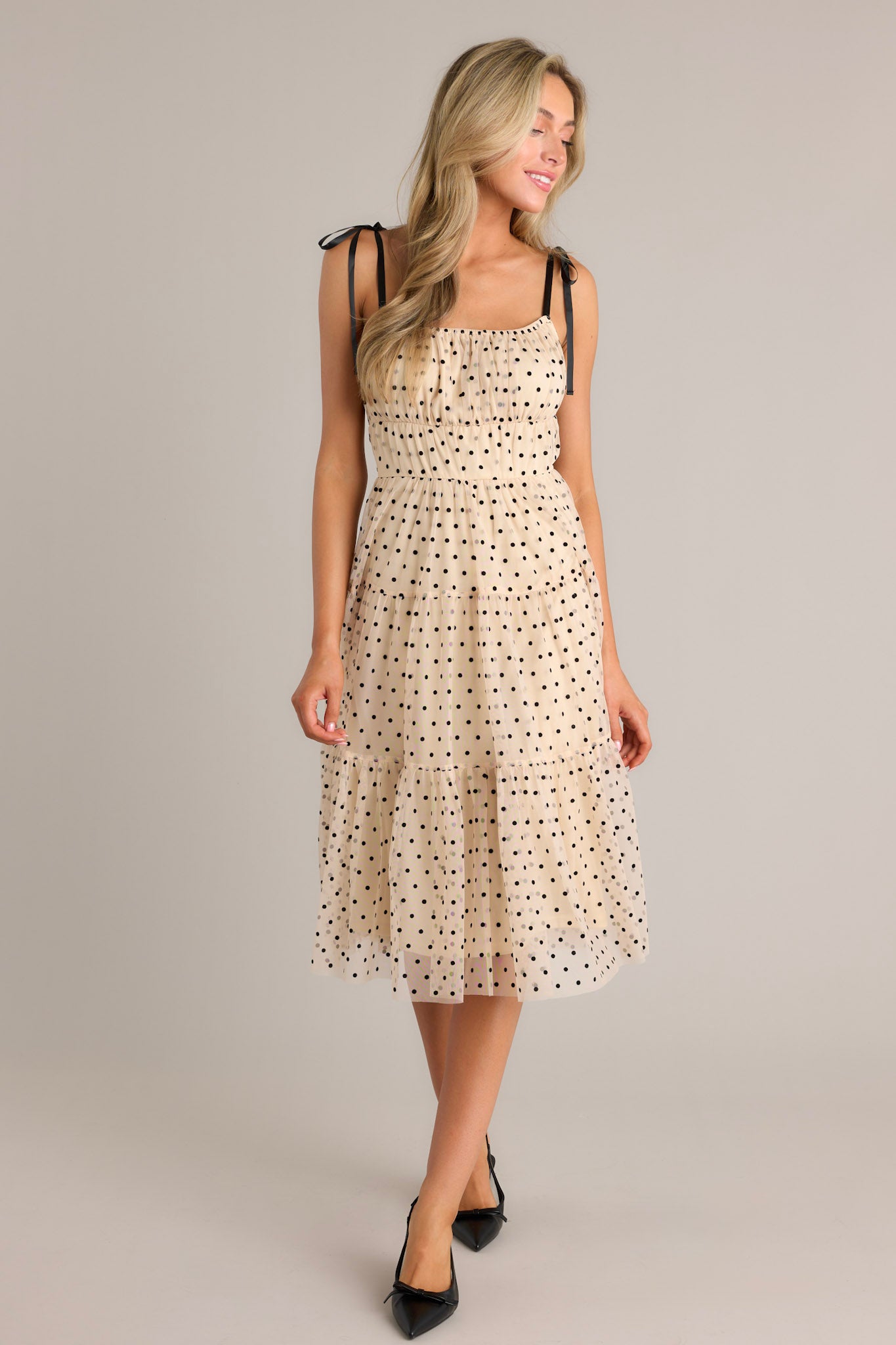 Front angled view of a beige midi dress featuring a square neckline, thin self-tie straps, an elastic waistline, tiers, black polka dots, and tulle