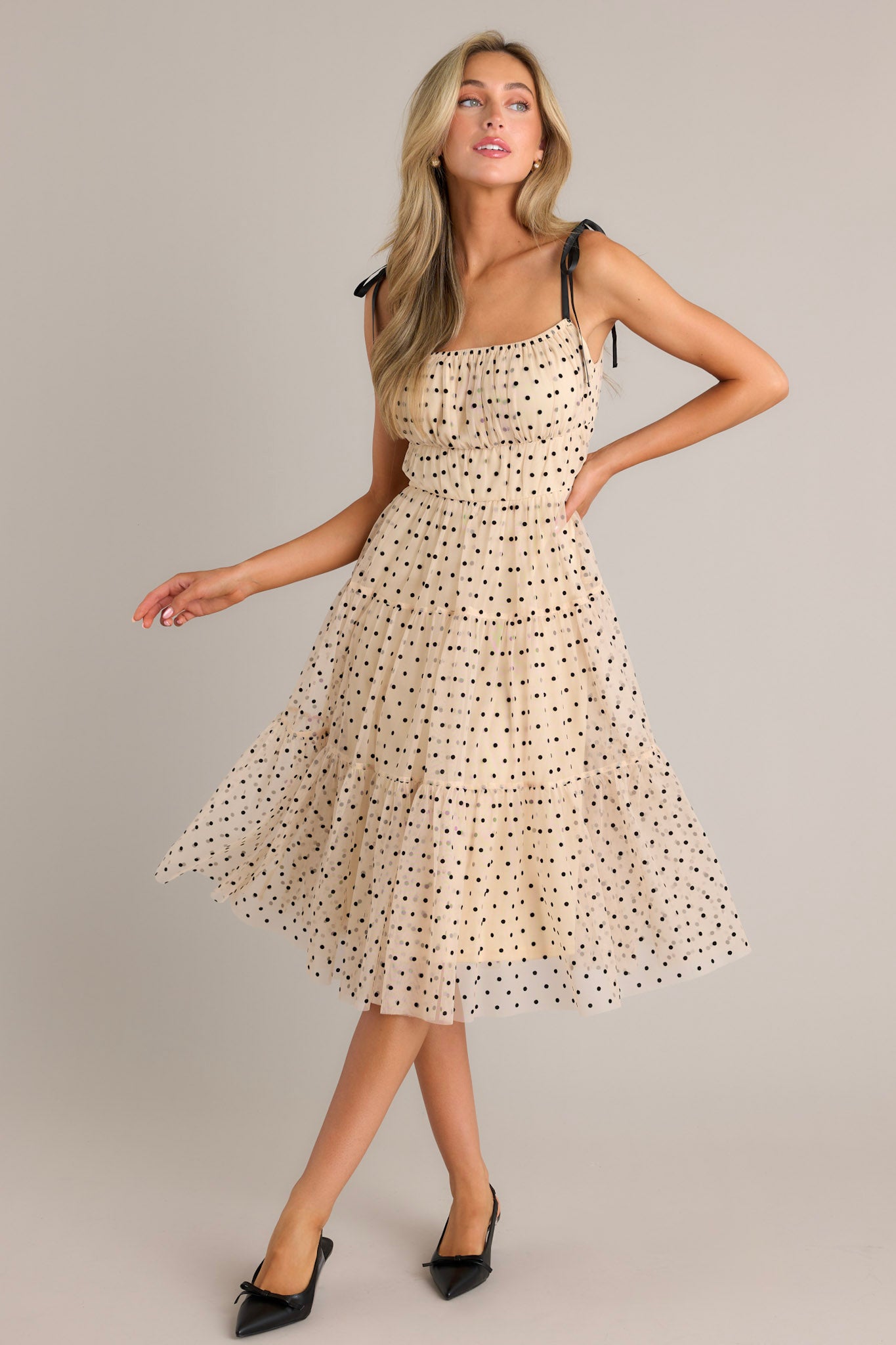Front view of a beige midi dress featuring a square neckline, thin self-tie straps, an elastic waistline, tiers, black polka dots, and tulle.