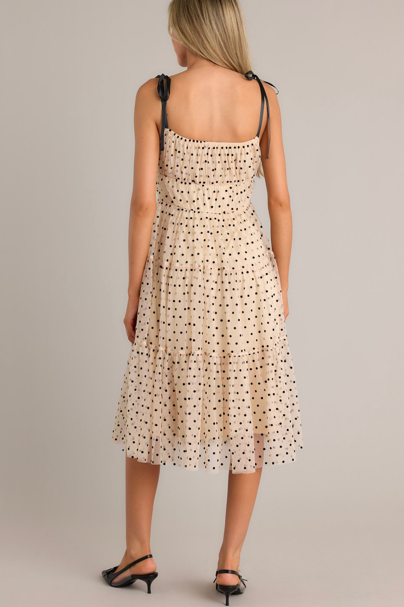 Back view of a beige midi dress highlighting the thin self-tie straps, tiers, black polka dots, and tulle.