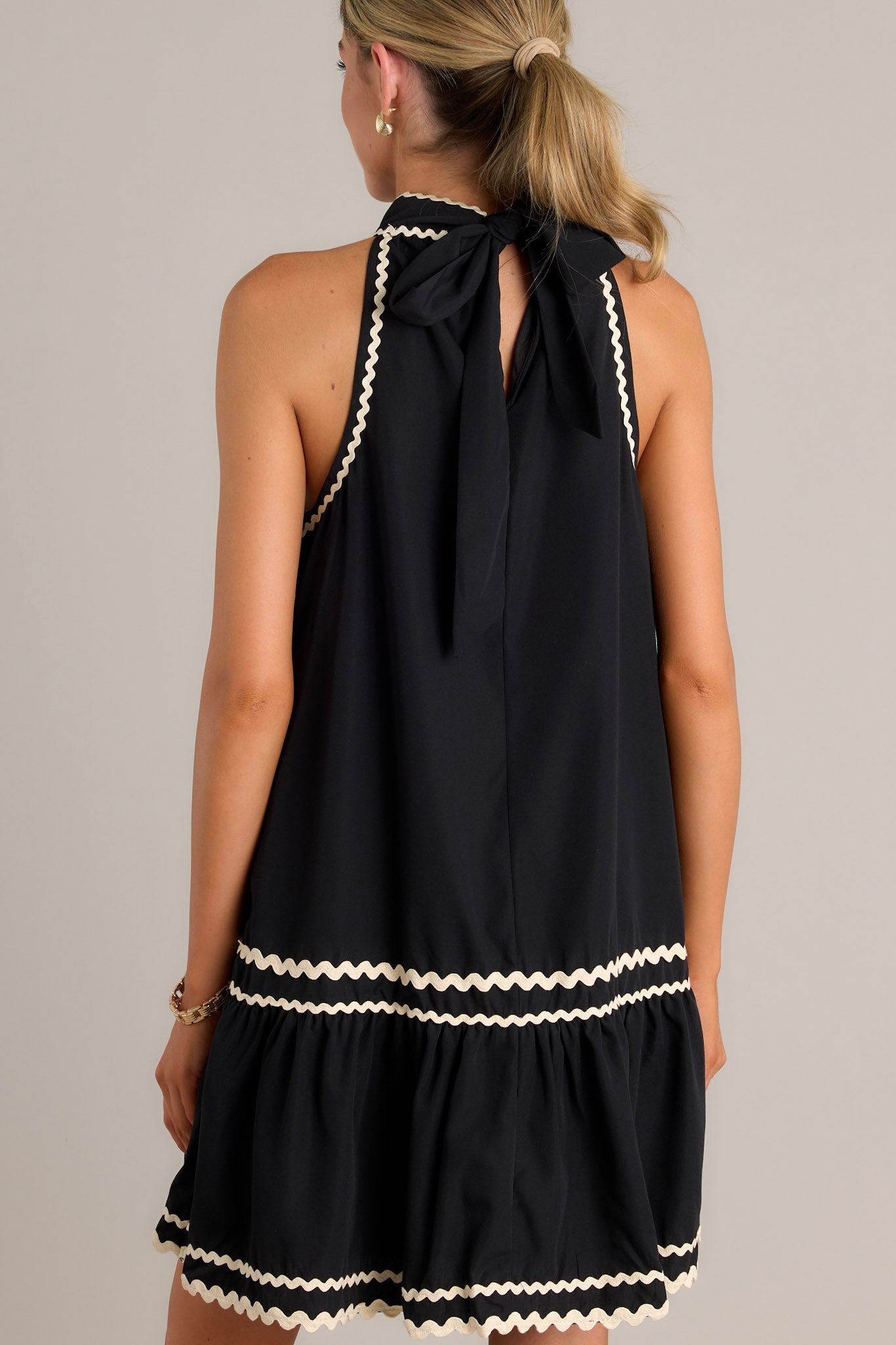 Back view of a black midi dress highlighting the high self-tie neckline, keyhole cutout, ivory ricrac detailing, and sleeveless design.