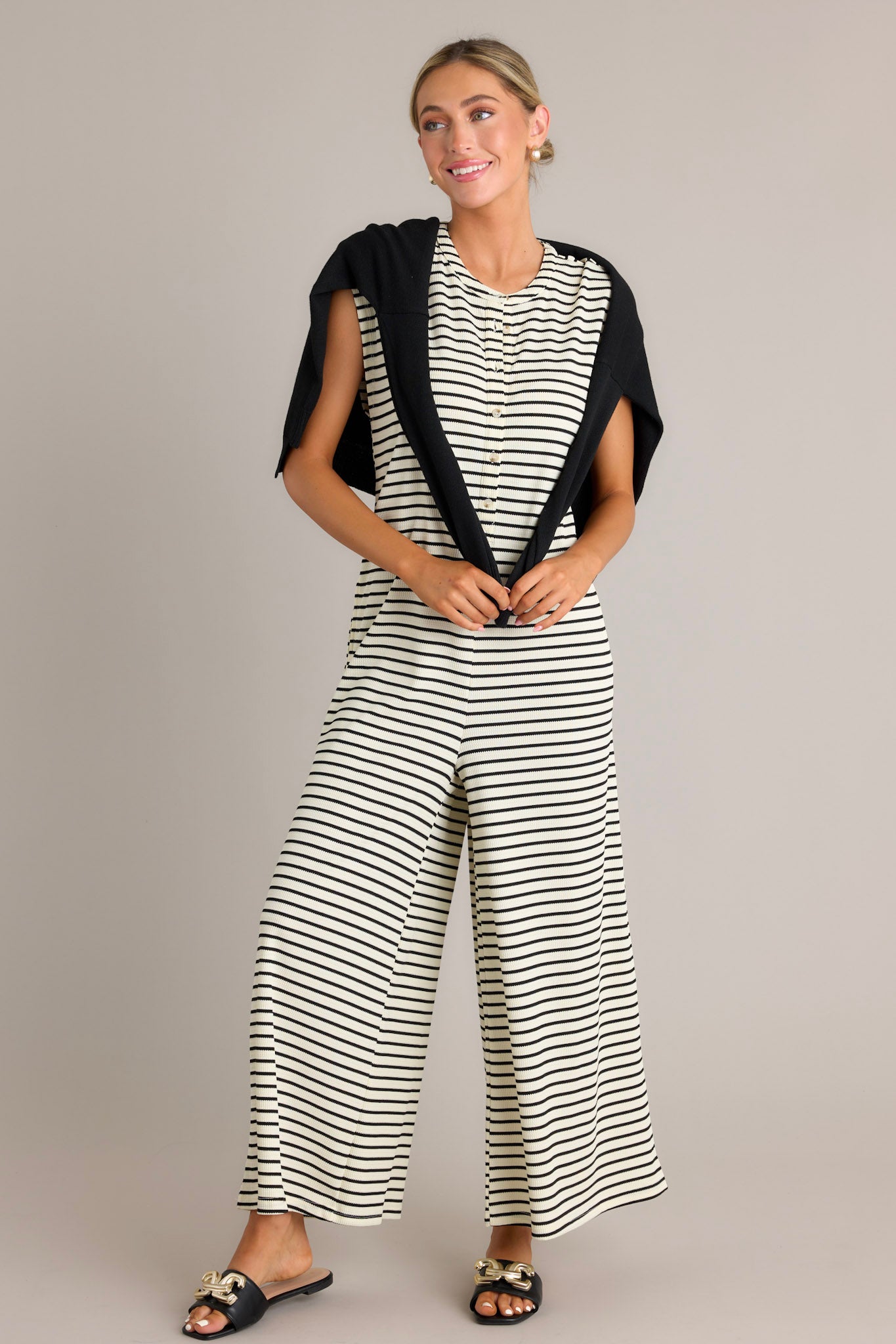 Full length view of a black striped jumpsuit with a rounded neckline, functional button front bodice, functional hip pockets, a textured material, and a sleeveless design