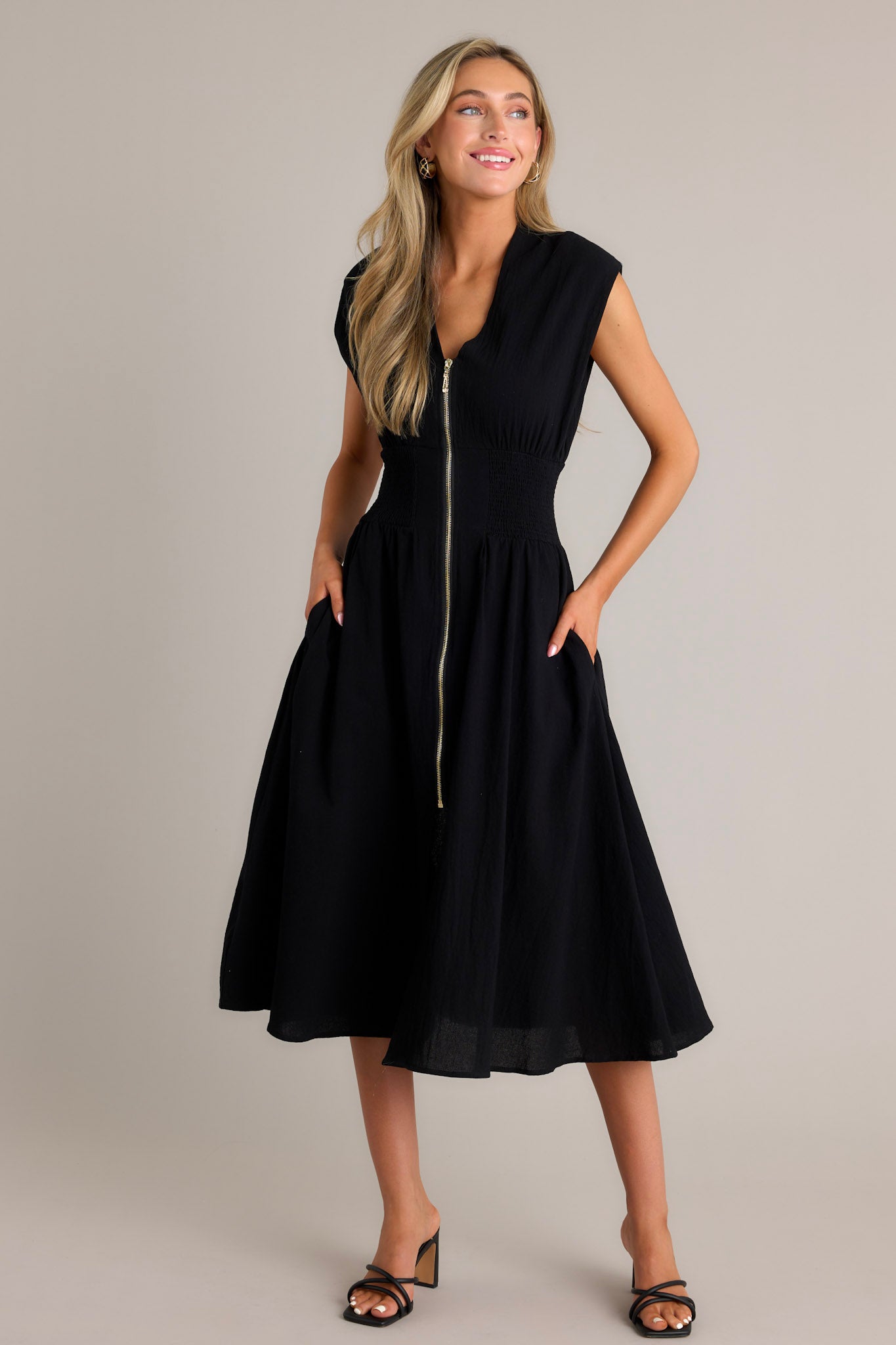 Full length view of a black midi dress with a deep v-neckline, shoulder padding, a functional zipper front, a fully smocked waist, functional pockets, and a front slit