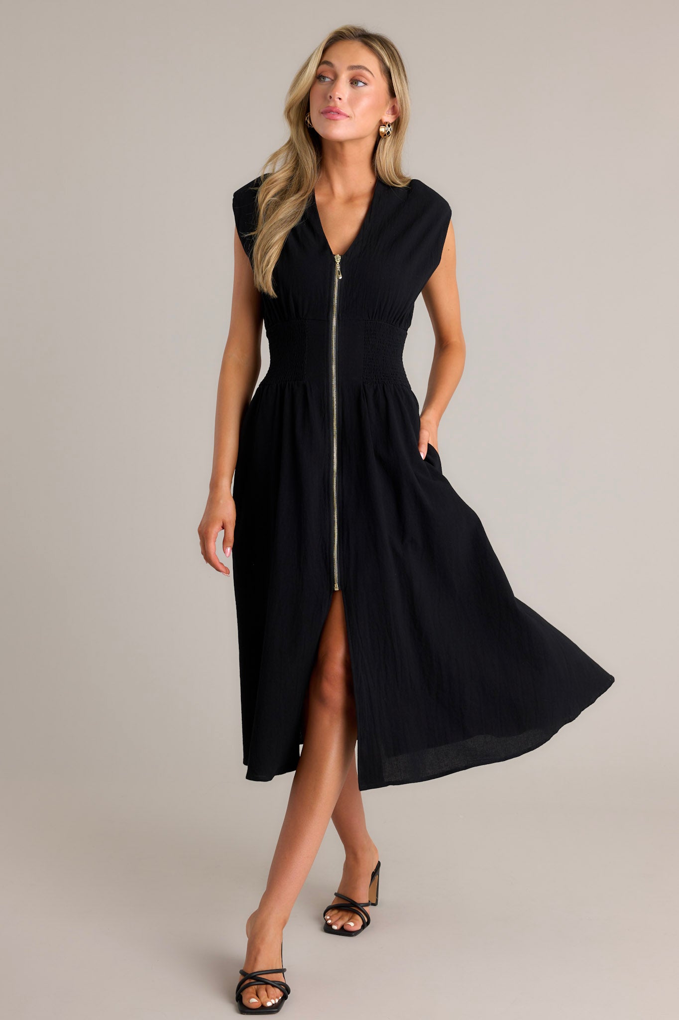 Action shot of a black midi dress displaying the fit and movement, highlighting the deep v-neckline, shoulder padding, functional zipper front, fully smocked waist, functional pockets, and front slit.
