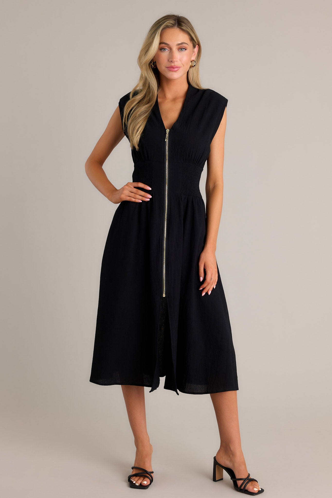 Front view of a black midi dress featuring a deep v-neckline, shoulder padding, a functional zipper front, a fully smocked waist, functional pockets, and a front slit.