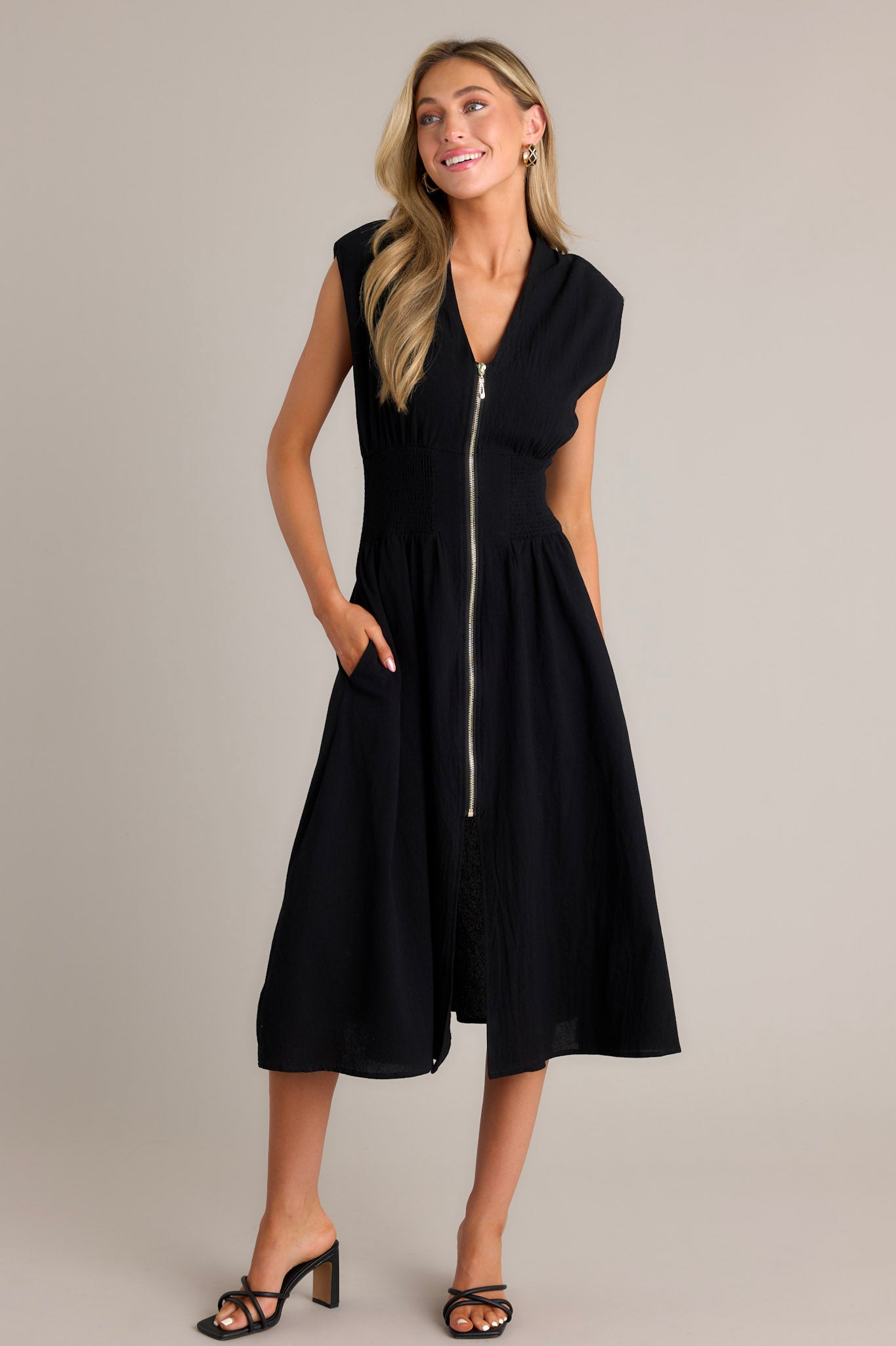 Side view of a black midi dress showcasing the deep v-neckline, shoulder padding, functional zipper front, fully smocked waist, functional pockets, and front slit.