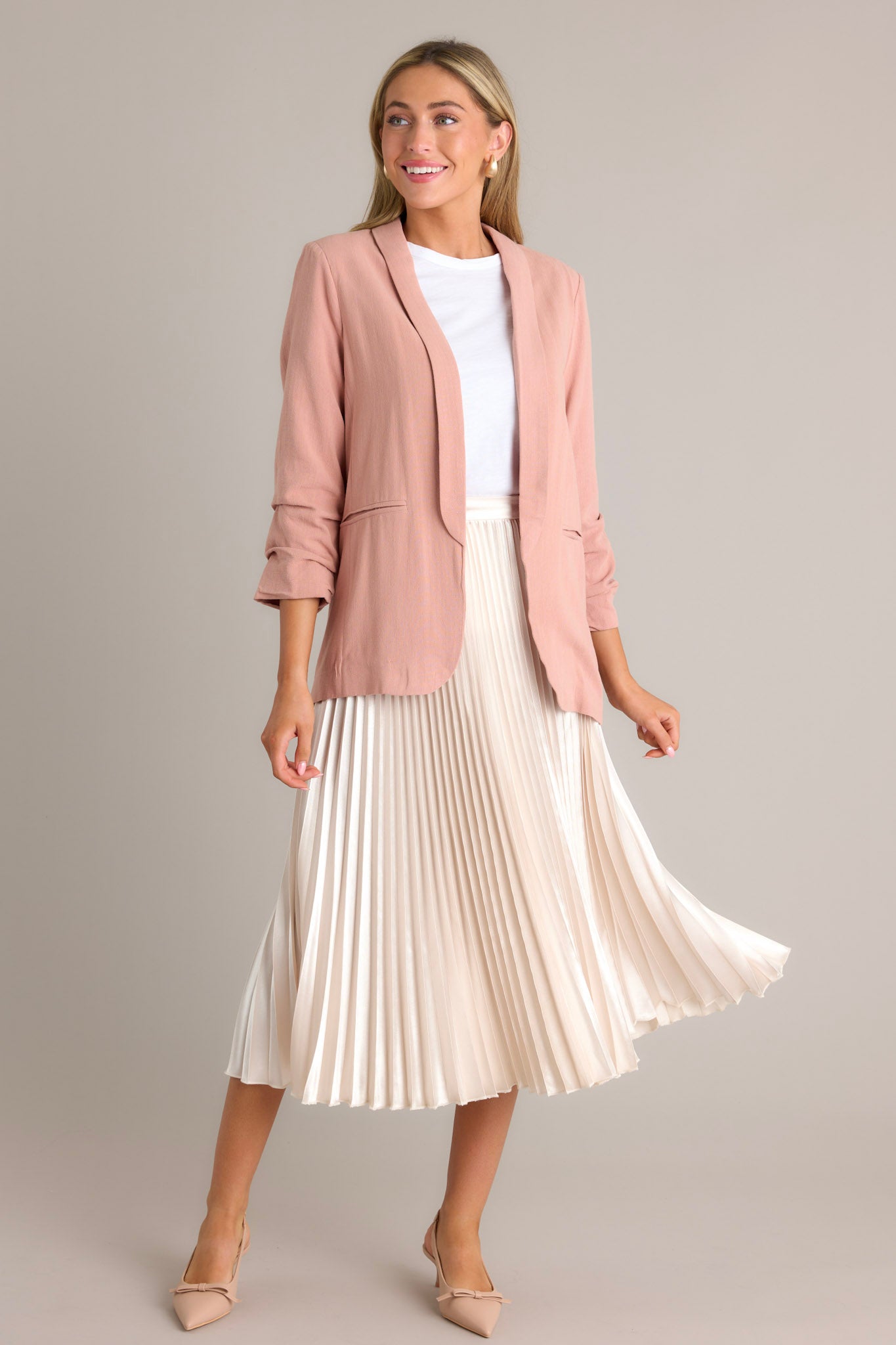 Angled full body view of this dusty rose blazer featuring a folded neckline, shoulder padding, faux pockets, and gathering in the quarter-length sleeves.