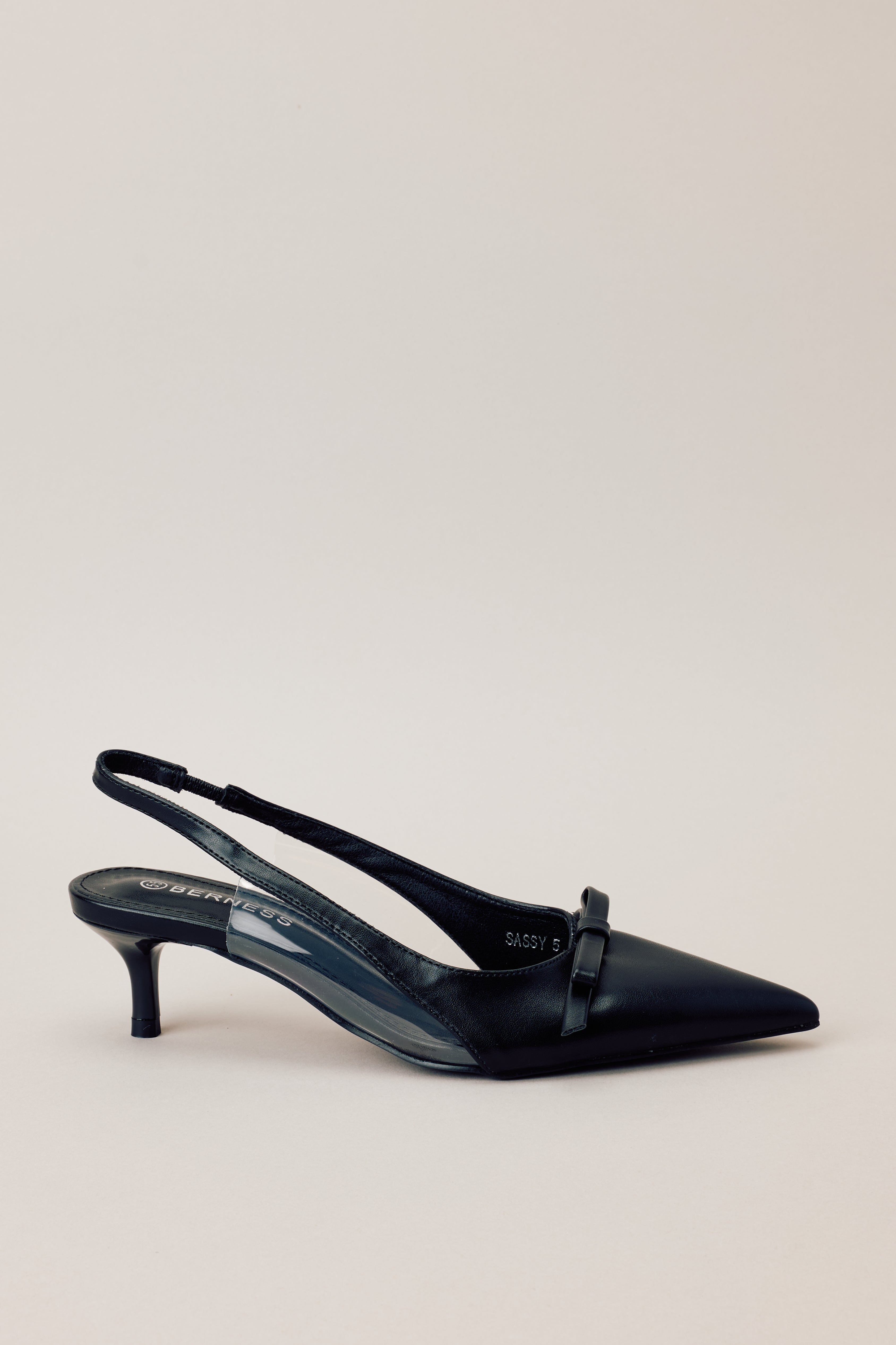 Side view of black kitten heels showcasing the pointed closed toe, delicate bow detailing, strap around the back of the foot, clear siding for extra support, and the short heel.