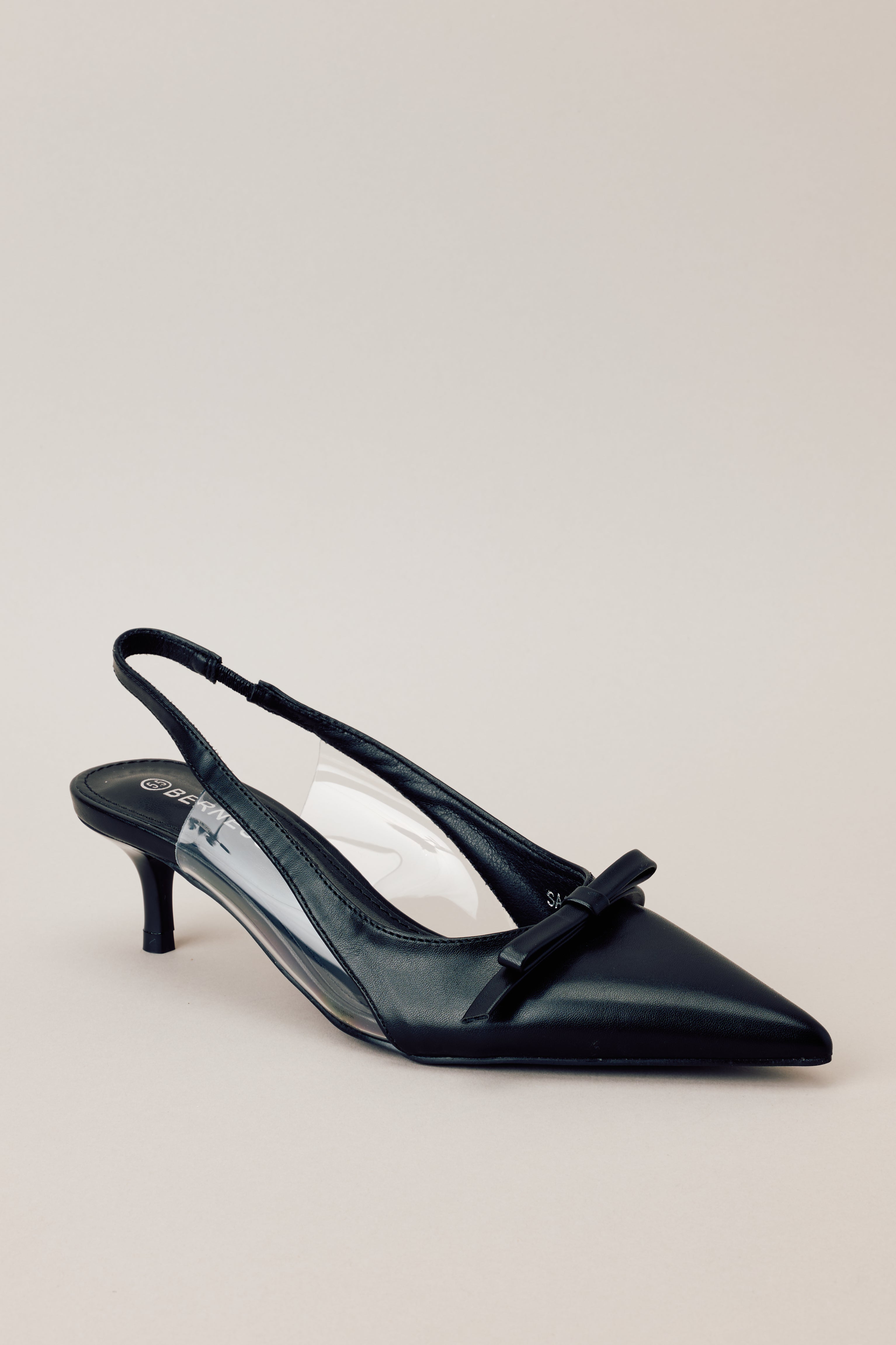 Front angled view of black kitten heels featuring a pointed closed toe, delicate bow detailing, a strap around the back of the foot, clear siding for extra support, and an extremely short heel