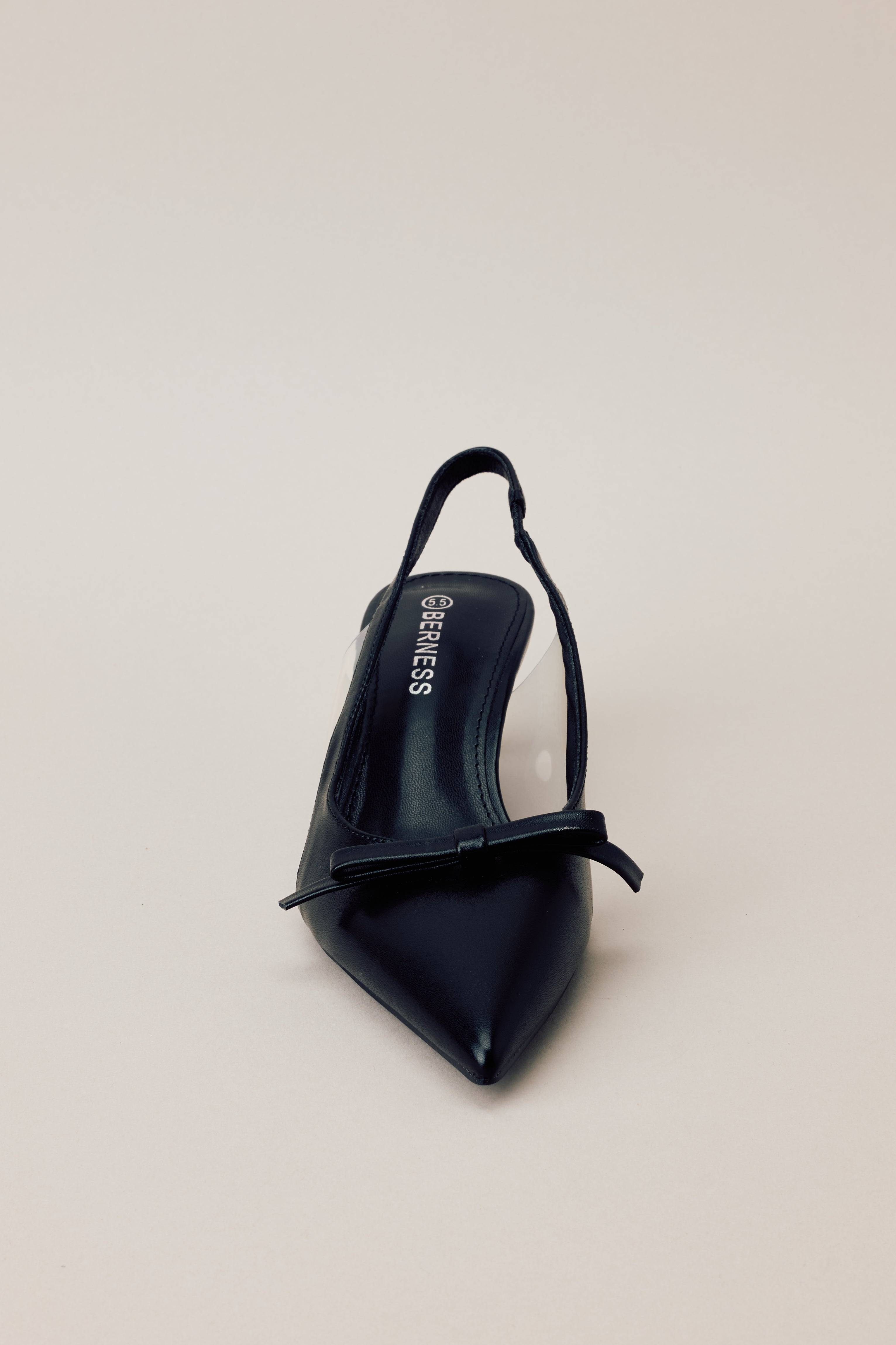 Front view of black kitten heels featuring a pointed closed toe, delicate bow detailing, a strap around the back of the foot, clear siding for extra support, and an extremely short heel.