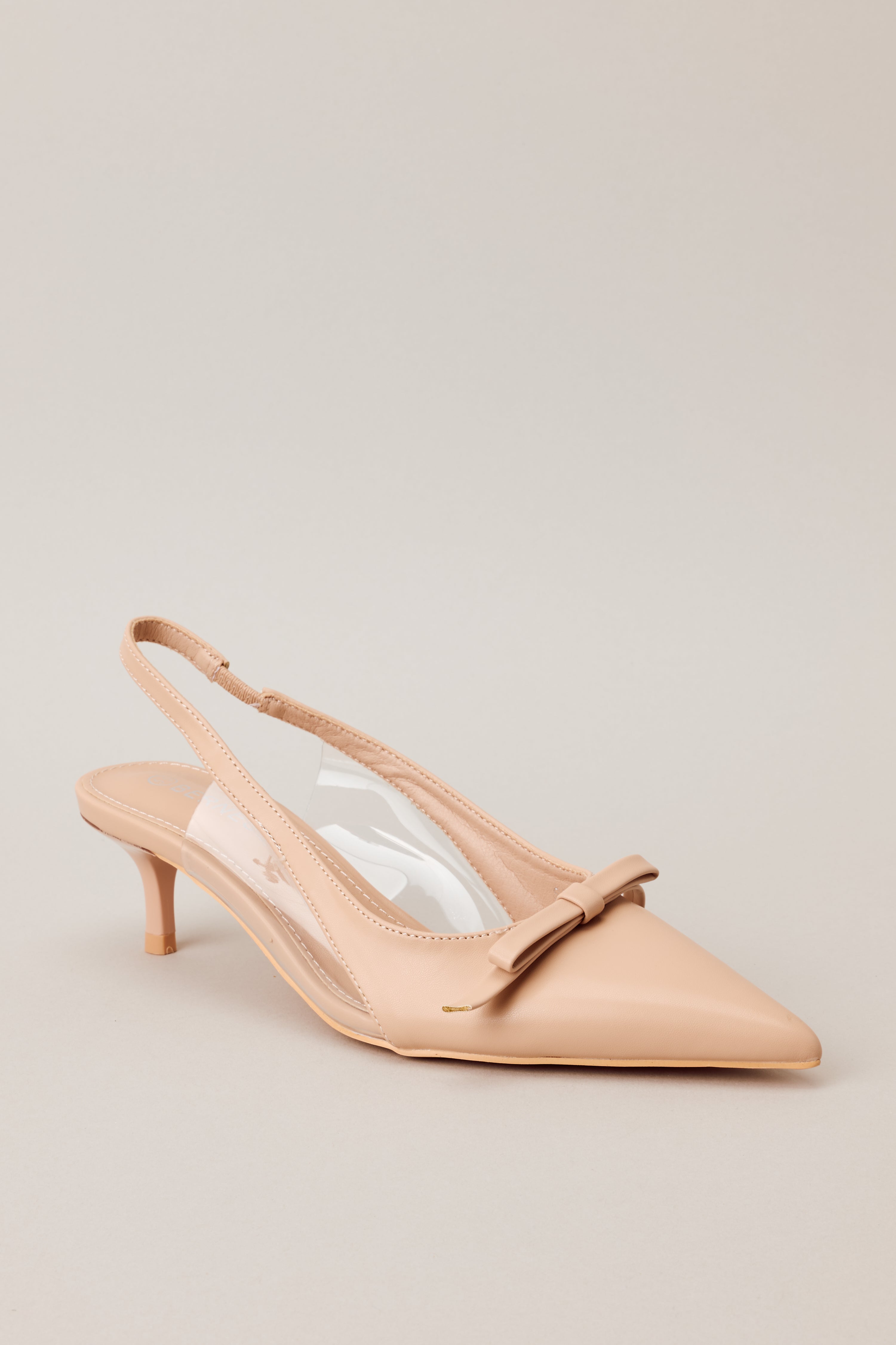 Close-up Front angled view of beige kitten heels featuring a pointed closed toe, delicate bow detailing, a strap around the back of the foot, clear siding for extra support, and an extremely short heel
