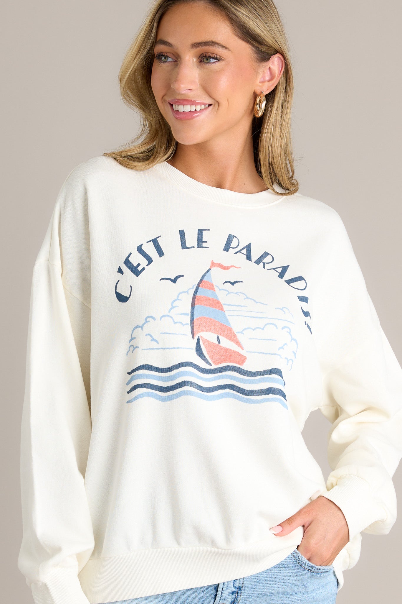 Front angled view of a white sweatshirt featuring a crew neckline, a sailboat themed graphic, ribbed cuffed long sleeves, and a ribbed hemline