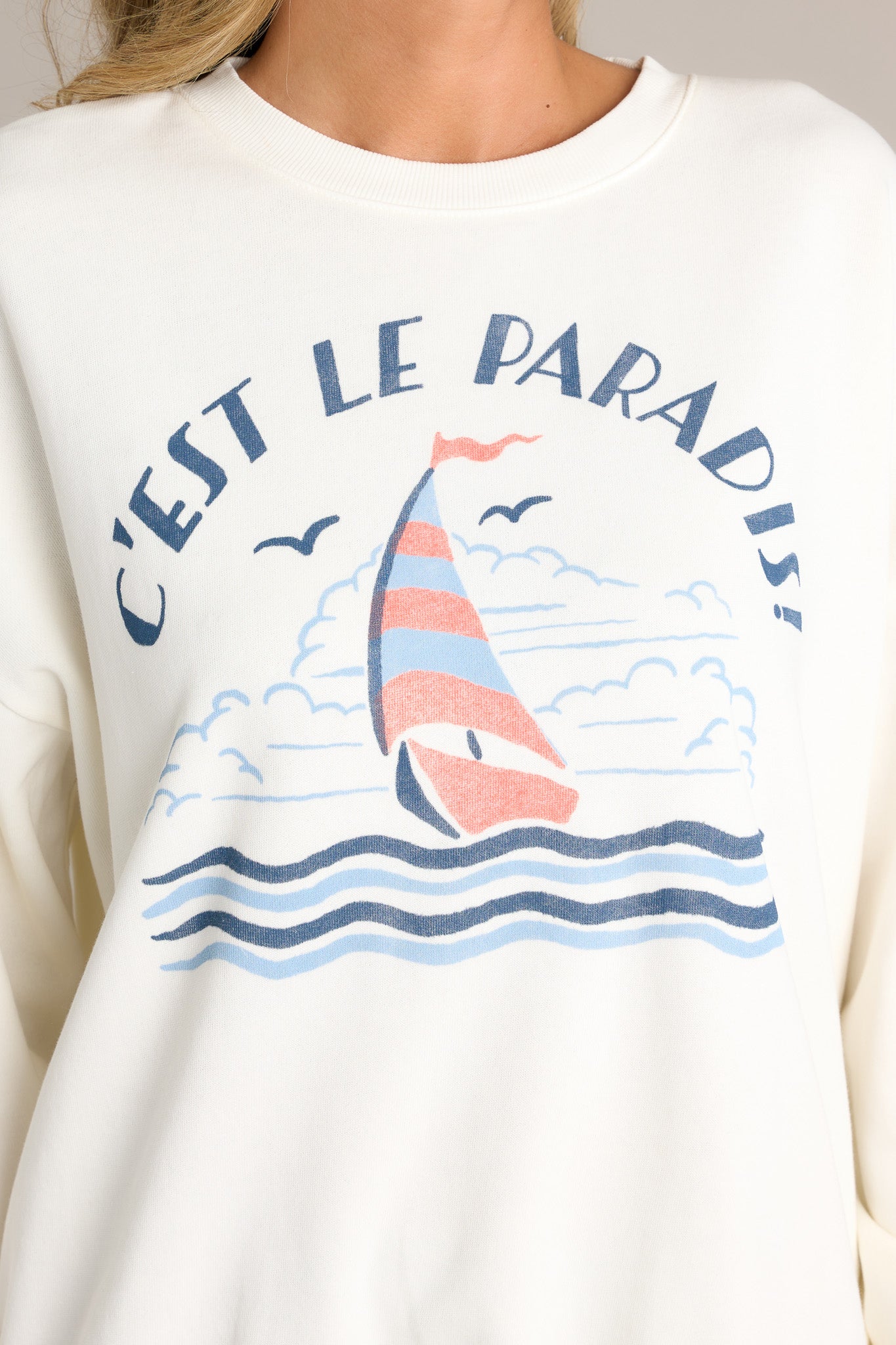 Close-up of the white sweatshirt showing the crew neckline, sailboat themed graphic, and ribbed cuffed long sleeves.