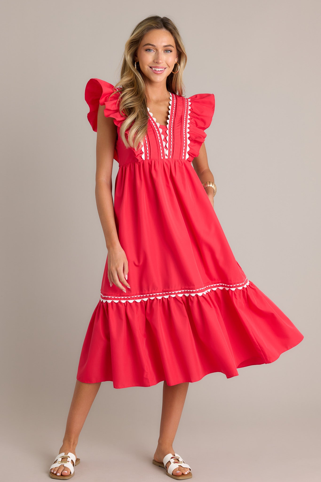 This red midi dress features a v-neckline, a discrete side zipper, scalloped & ruffled bodice detailing, functional hip pockets, and short flutter sleeves.