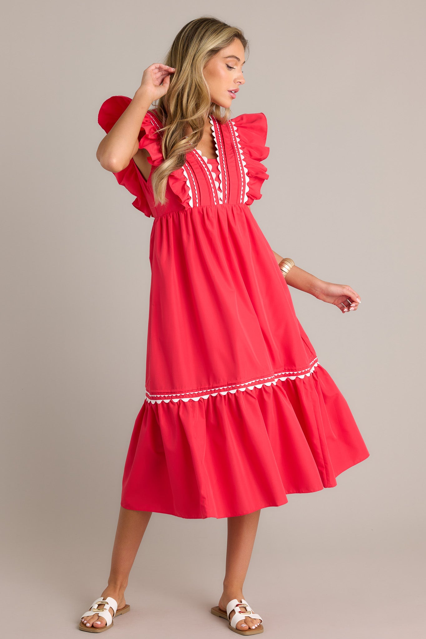 Full length view of a red midi dress with a v-neckline, a discrete side zipper, scalloped and ruffled bodice detailing, functional hip pockets, and short flutter sleeves