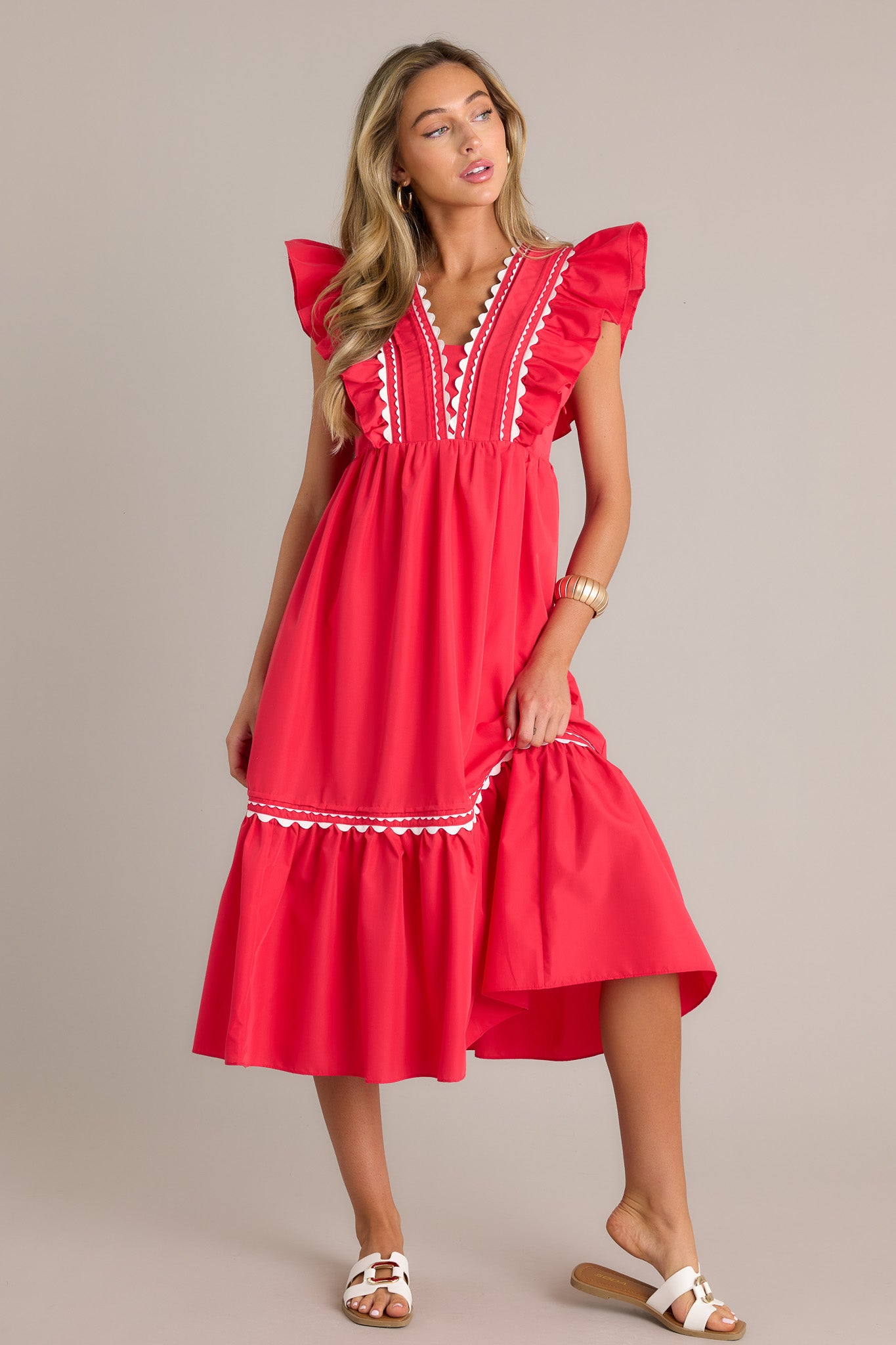 Front angled view of a red midi dress featuring a v-neckline, a discrete side zipper, scalloped and ruffled bodice detailing, functional hip pockets, and short flutter sleeves