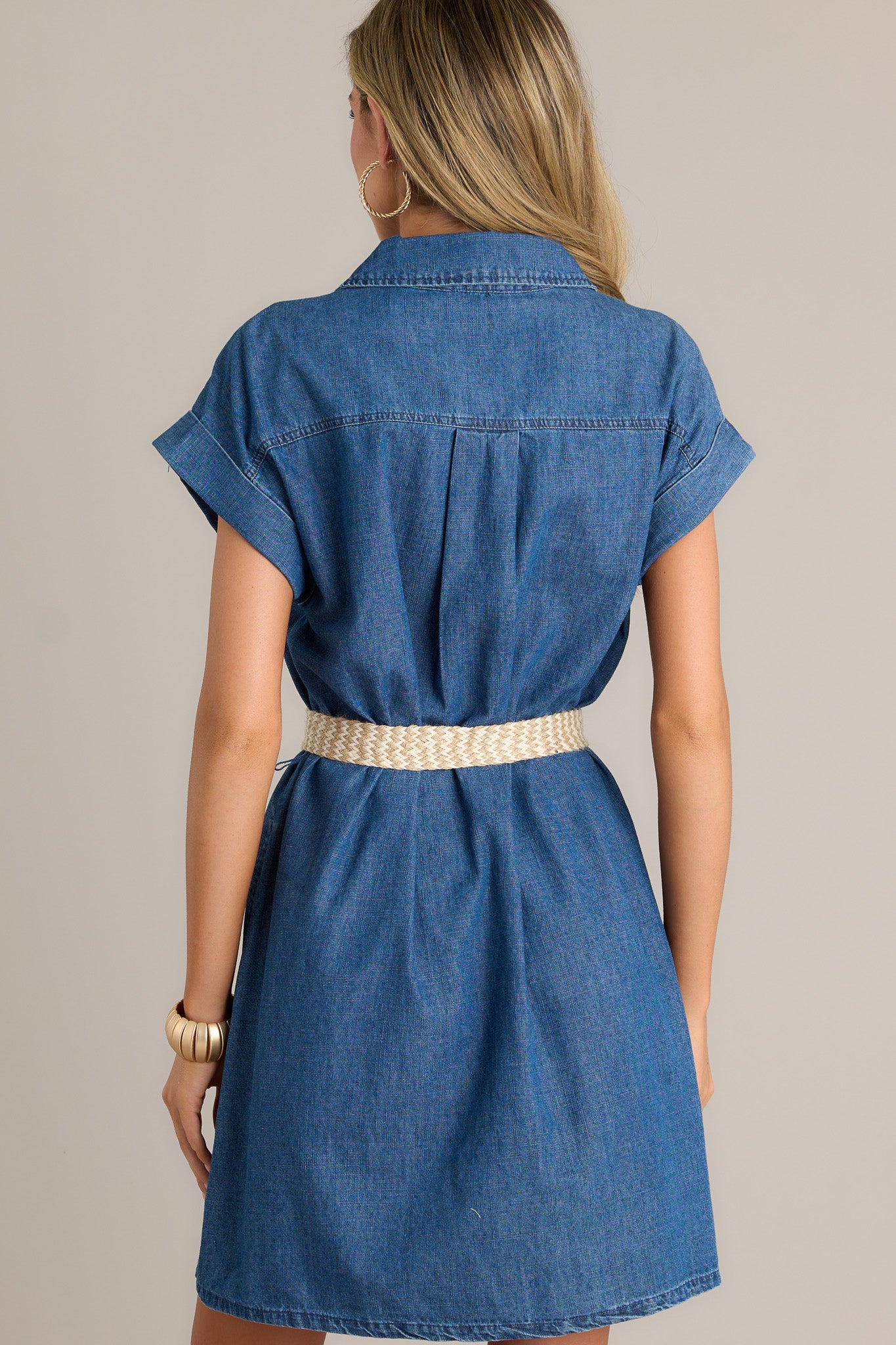 Back view of a dark denim mini dress highlighting the pull-through belt, cuffed short sleeves, and overall fit.