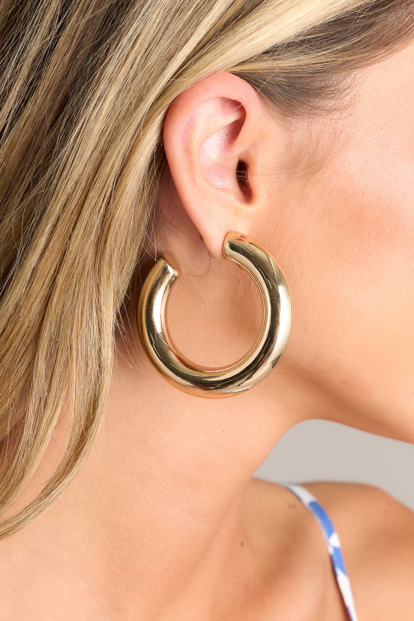 Close-up of a large gold hoop earring with an open circular design. The earring features a smooth, polished surface that reflects light.