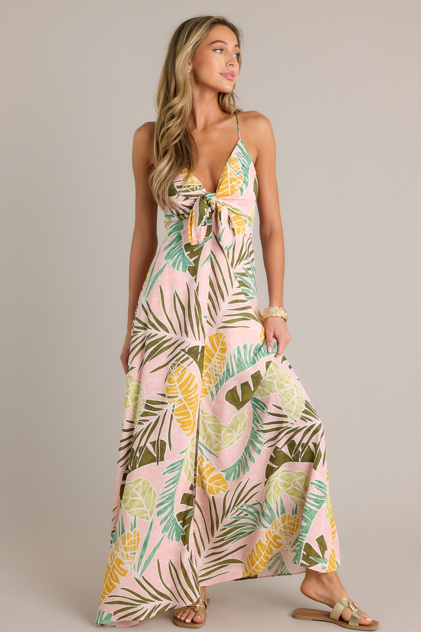 Angled front view of a light pink maxi dress showcasing the v-neckline, thin adjustable self-tie straps, self-tie knot in the bust, unique palm pattern, front slit, and flowing silhouette.