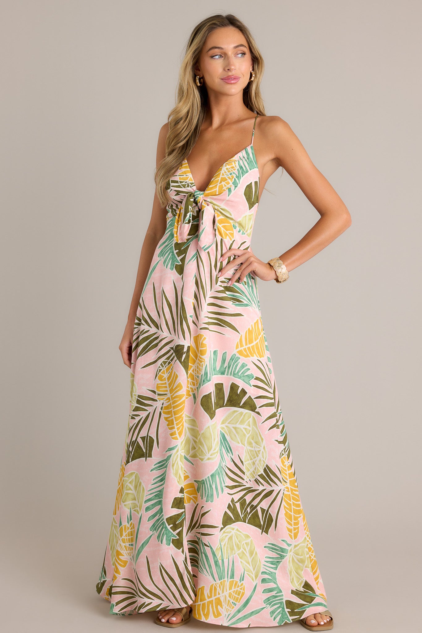 This light pink maxi dress features a v-neckline, thin adjustable self-tie straps, a low back, a discrete zipper, a self-tie knot in the bust, a unique palm pattern, a front slit, and a flowing silhouette.