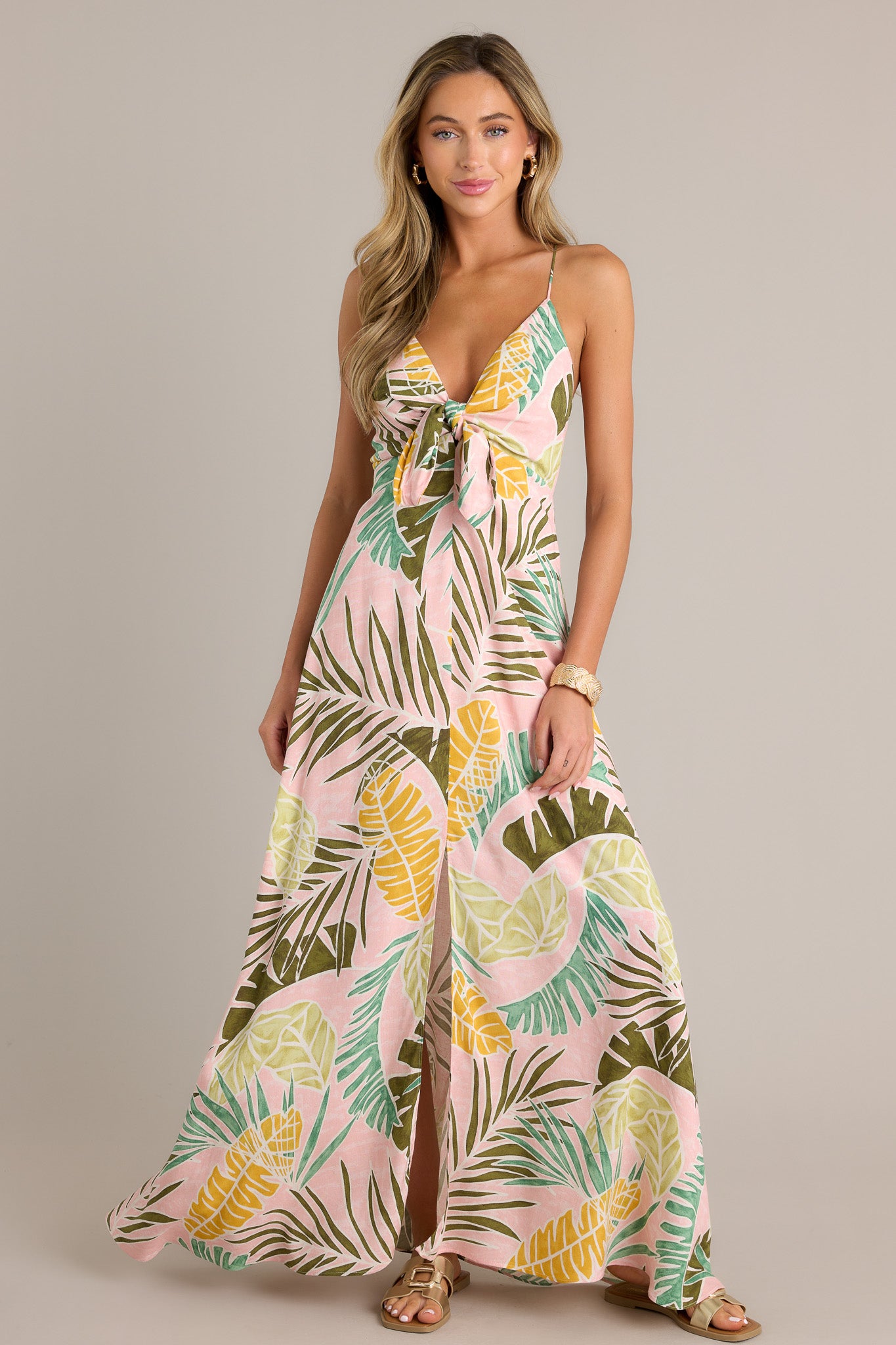 Full length view of a light pink maxi dress with a v-neckline, thin adjustable self-tie straps, a low back, a discrete zipper, a self-tie knot in the bust, a unique palm pattern, a front slit, and a flowing silhouette