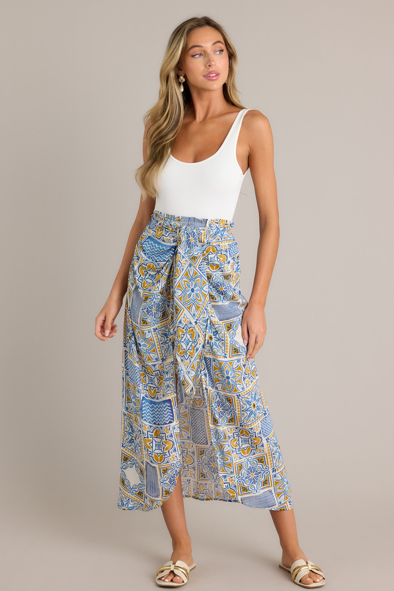 Front angled view of a blue maxi skirt featuring a high waisted design, an elastic waistband, a self-tie knot, an open front, a short skirt underneath, and a unique print