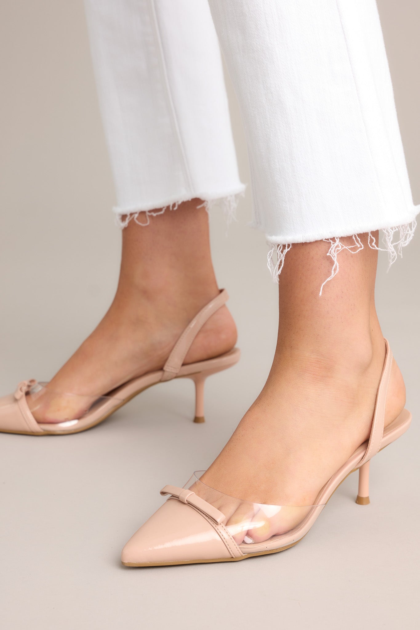 These beige kitten heels feature a pointed toe, delicate bow detailing, a strap around the back the foot, clear siding for extra support, and a short heel.