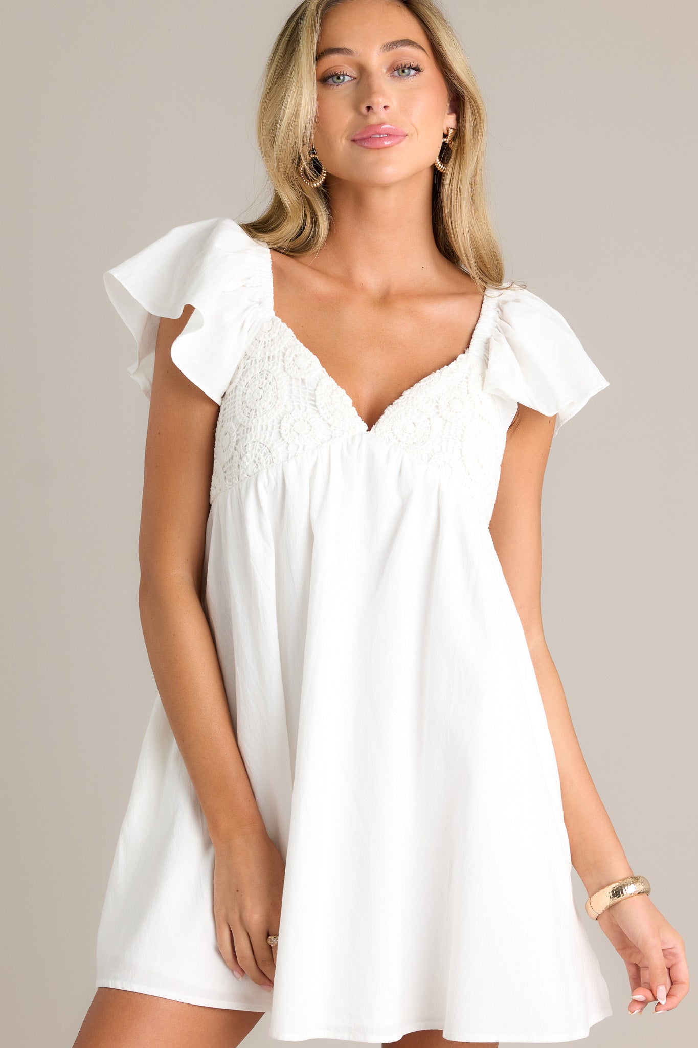 Front view of an ivory mini dress featuring a v-neckline, fully smocked back, unique knitted pattern on the bust, functional hip pockets, short flutter sleeves, and a flowing silhouette.