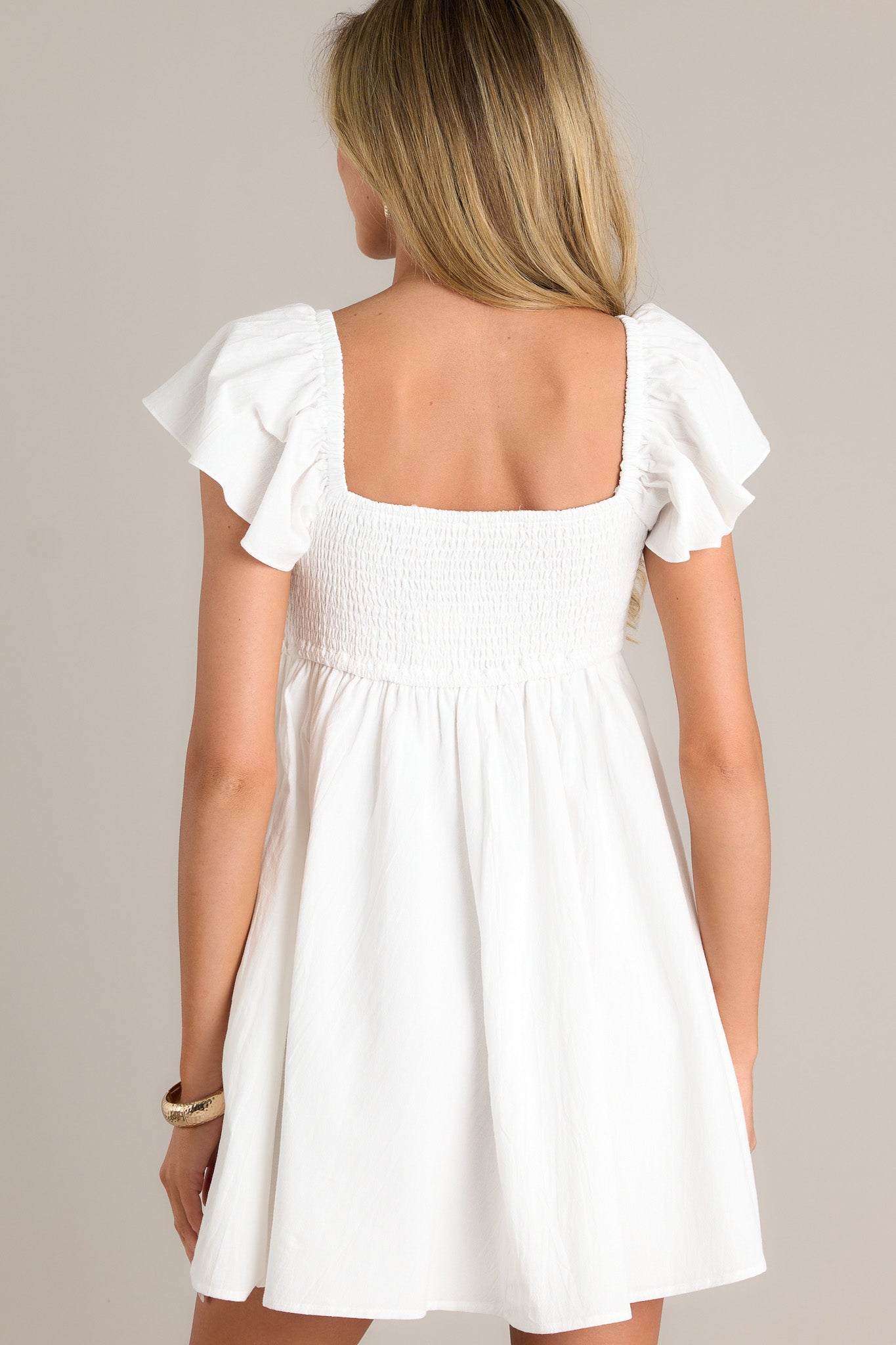 Back view of an ivory mini dress highlighting the fully smocked back, short flutter sleeves, and overall fit.